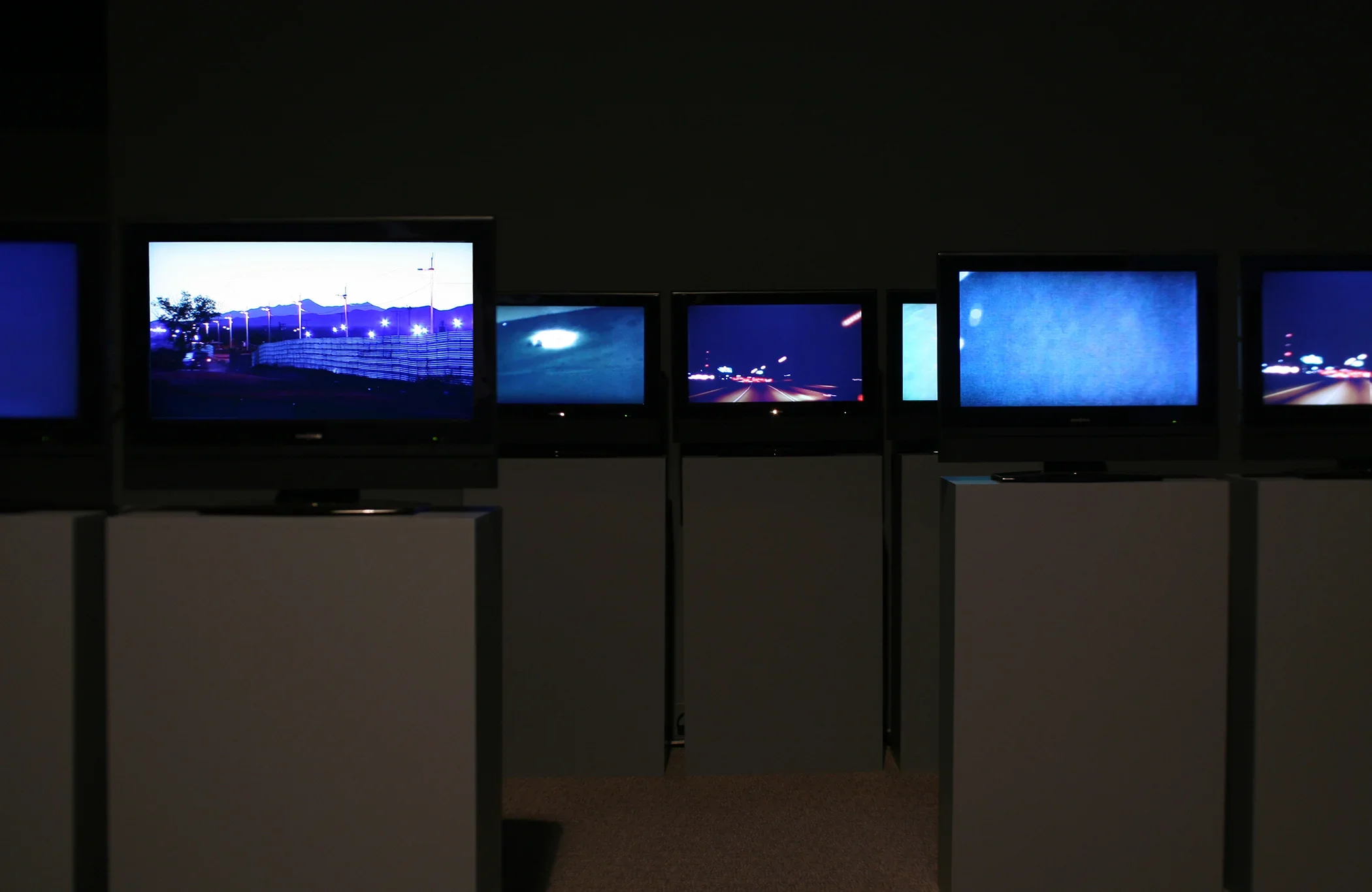 Many screens installed on podiums in a darkened gallery. 