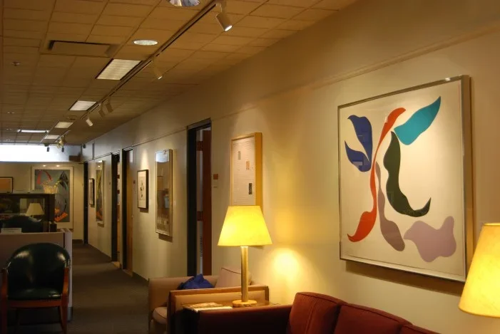 An entrance to a cosy office space with minimal abstract prints hung on the wall above a couch and chair with lamps beside.