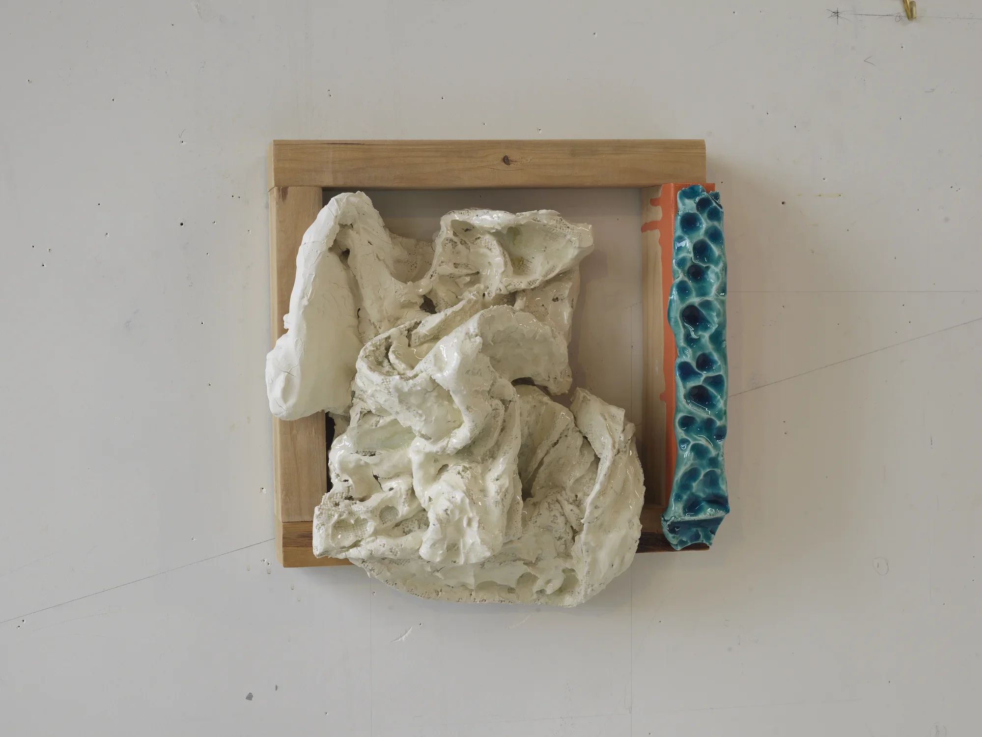 A hanging wall sculpture of white ceramic forms within a square wood frame partly covered in blue ceramic and red paint