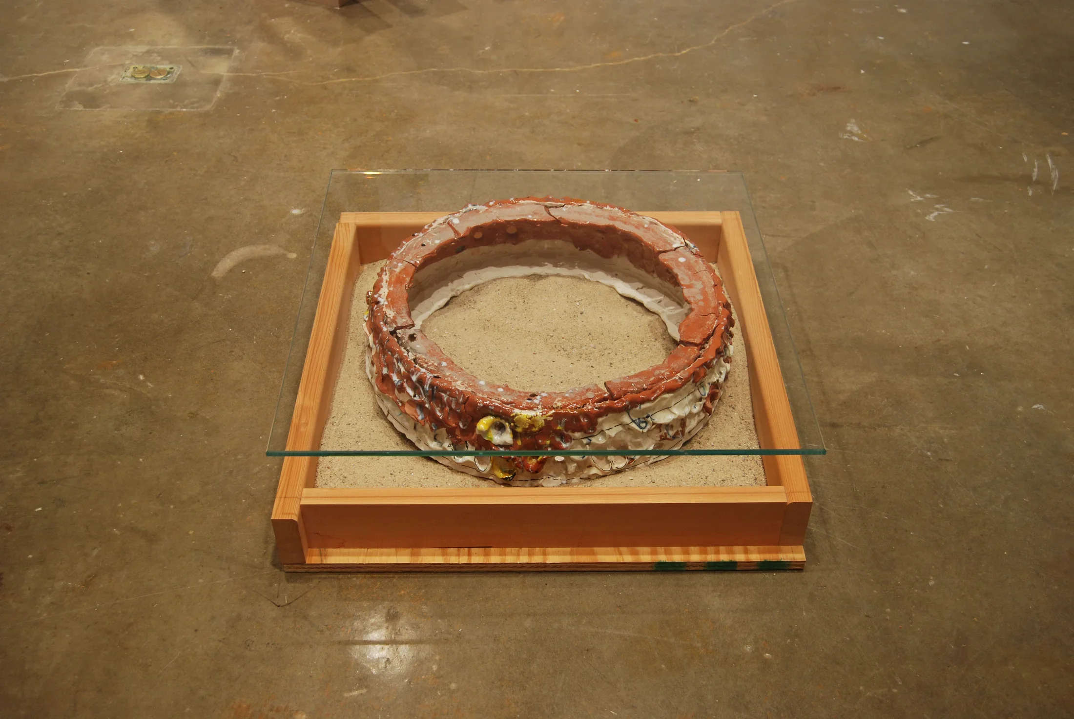 Close up of a floor sculpture with a square wood frame holding sand, a round ceramic shape, and a flat piece of glass on top