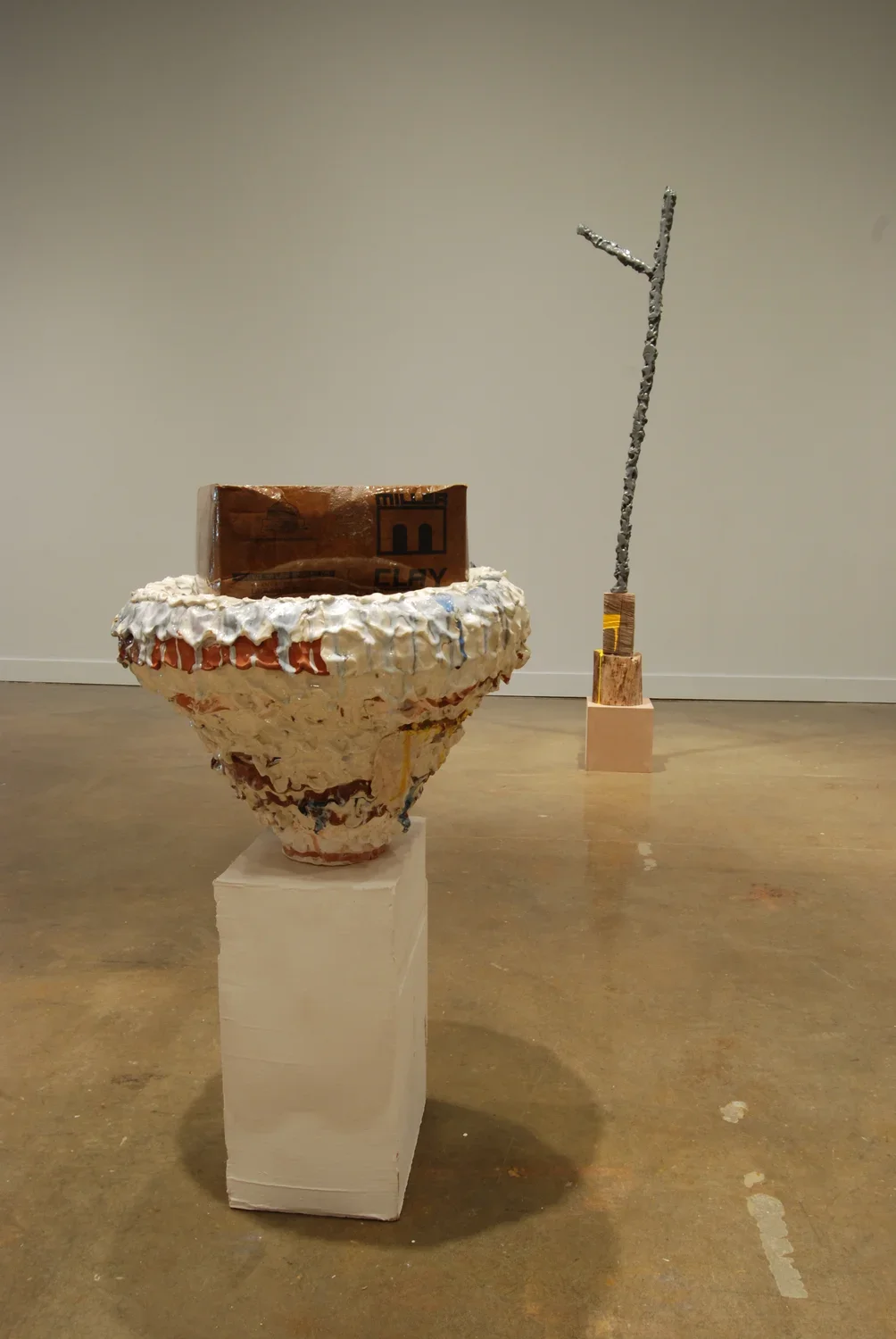 A round ceramic urn shape on a pedestal with a brown brick shape on top, and a standing branch like stick sculpture in back