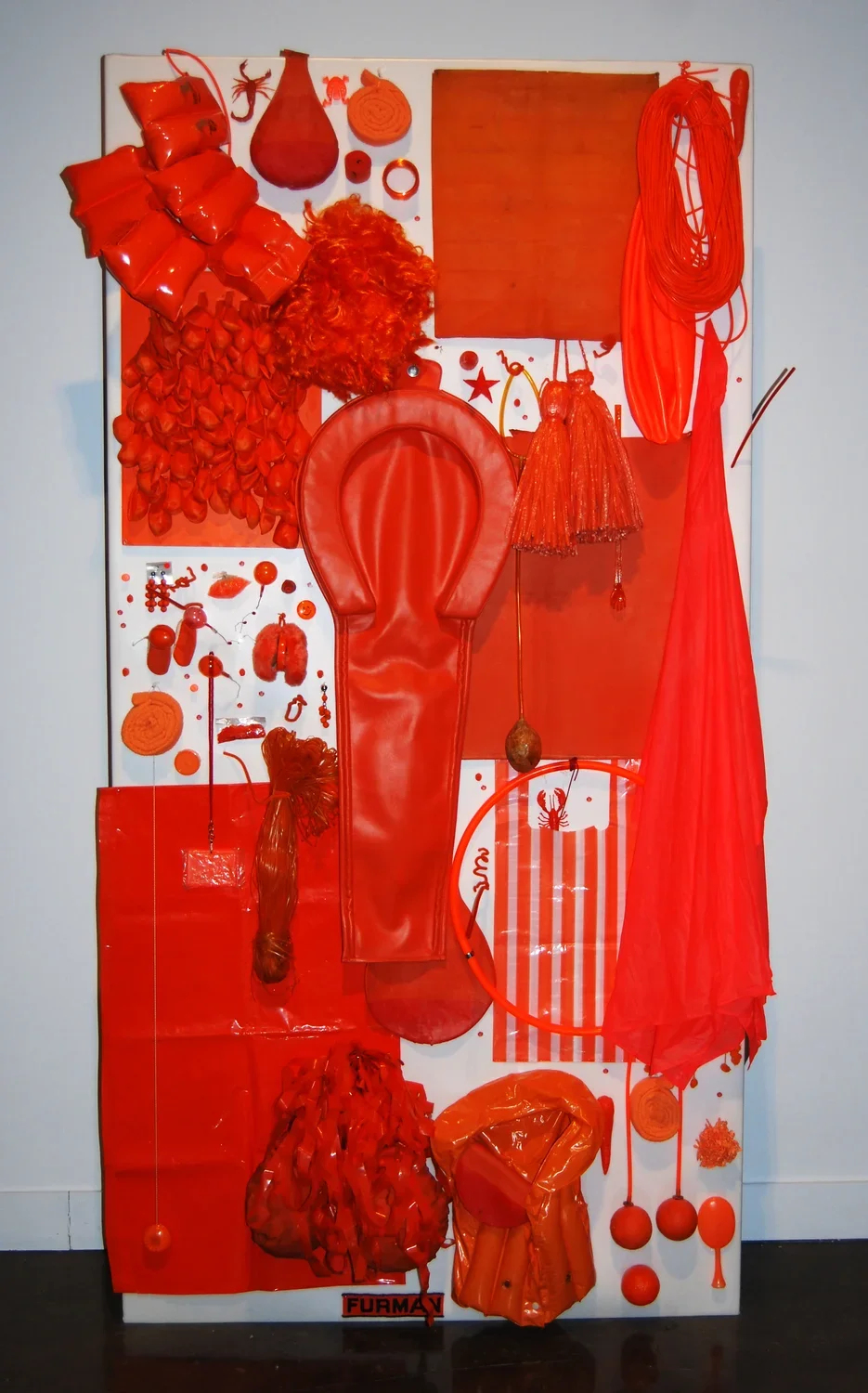 A standing white panel with bright orange objects attached; a large horseshoe, tassels, a round balloon shape, balls, stripes