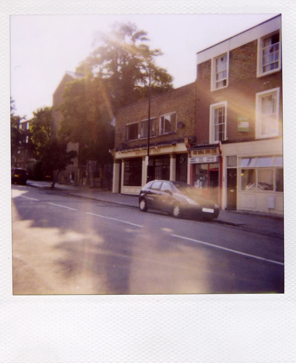 Streaks of warm light and faint reflections partly obscure the view of a streetscape with gray car, storefronts and trees.