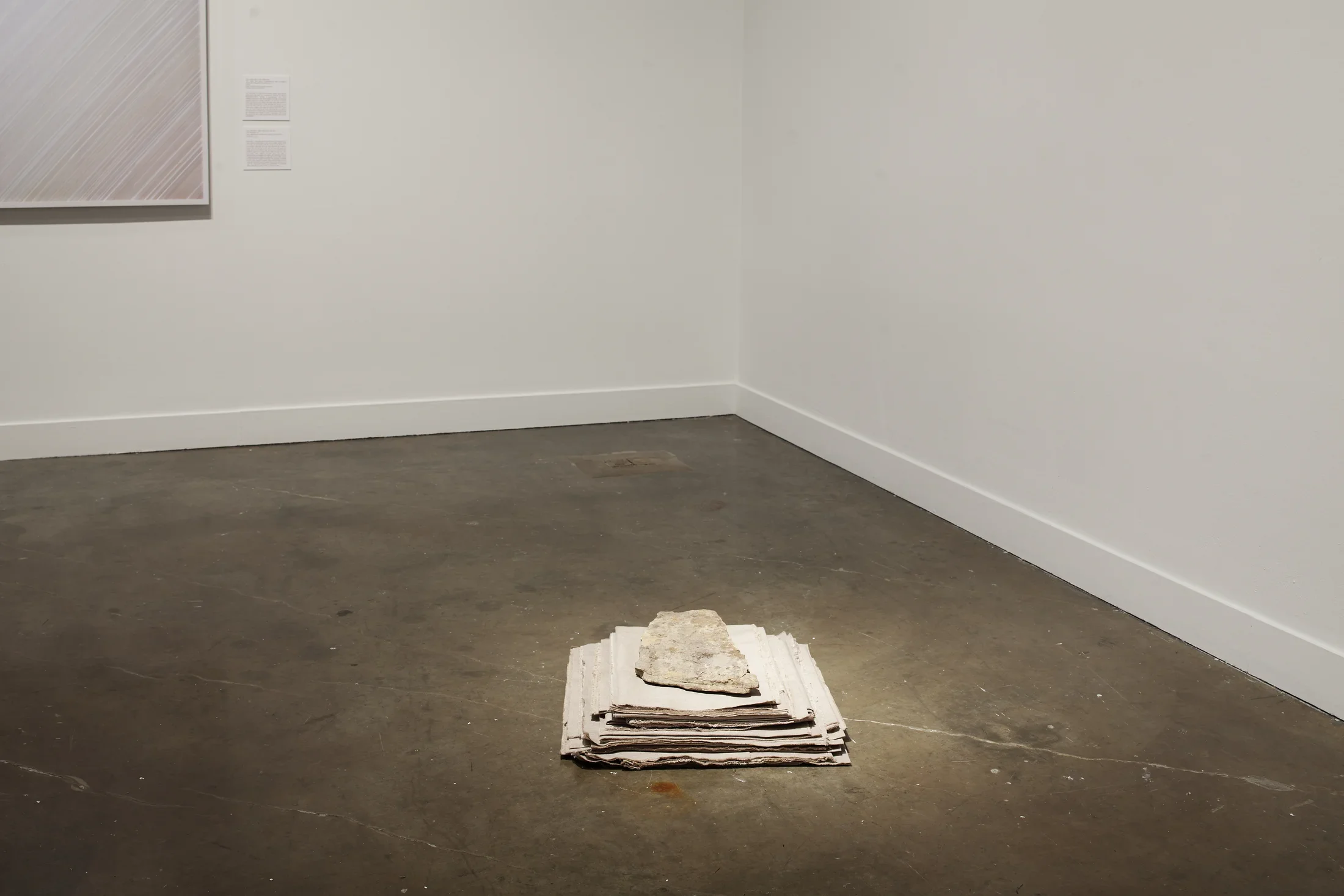 A pile of folded fabric, largest at the bottom, with a flat rock on top and placed on the concrete floor near a corner