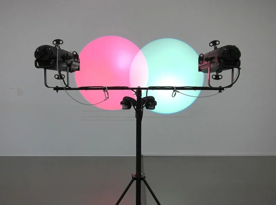 Two lamps on a stand, both at equal height, cast a pink and a green circle of light which overlap one another in the center.