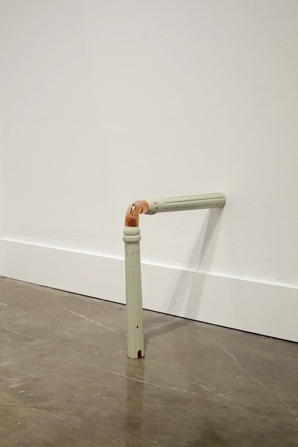 Two painted wooden chair legs, joined by an elbow of copper pipe, connect at the wall and floor at a 90 degree angle.