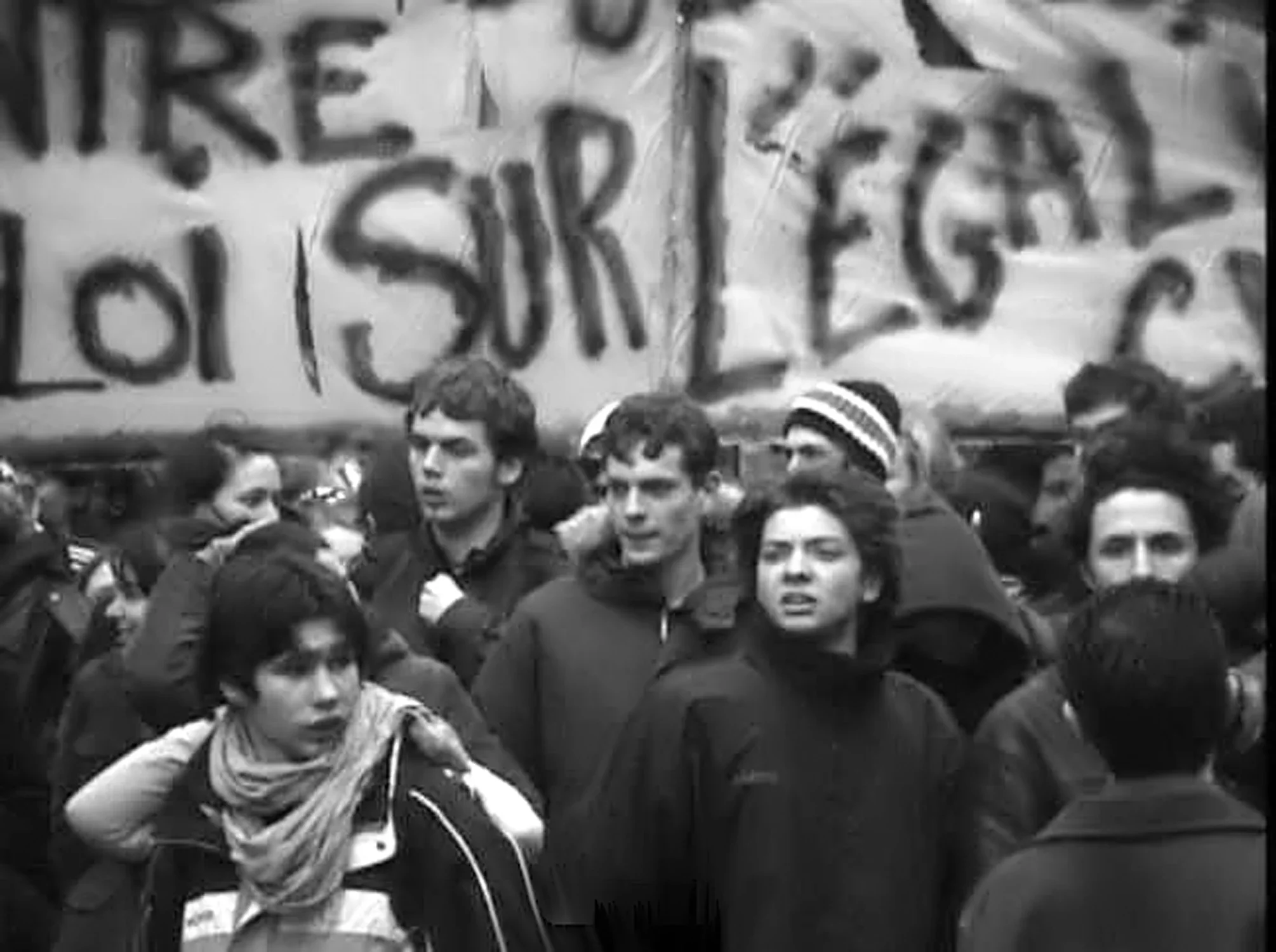 Black and white photo of several young people gathering in protest, a large handwritten sign filling the background.