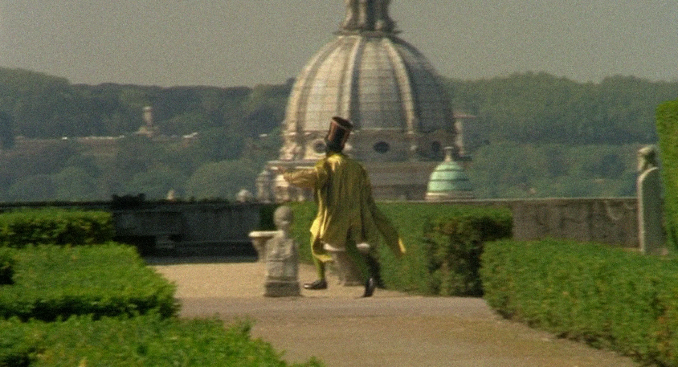 A man wearing a tunic and tall hat hurries away past hedgerows. An ornate white dome and mountains are seen in the background.