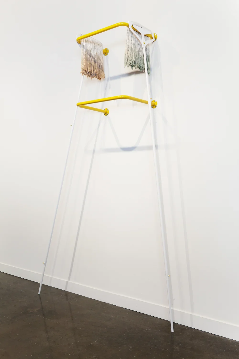 Two yellow railings mounted one over the other support 2 long white handled mops, heads draped over the sides of the top bar.