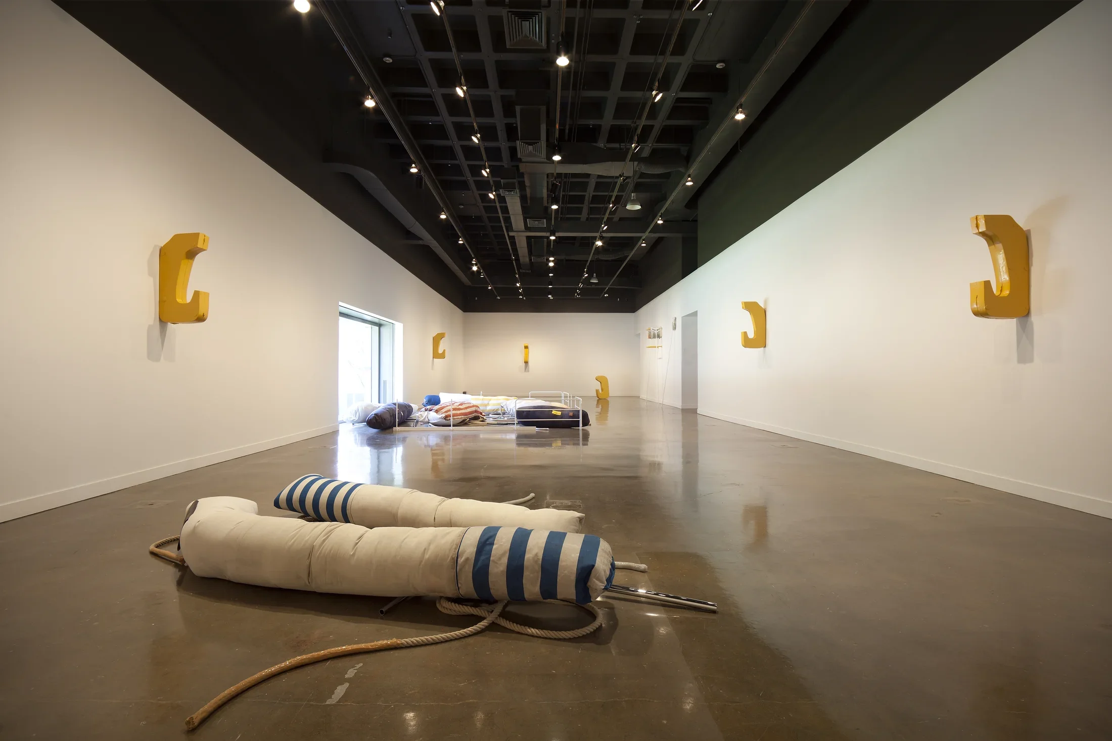 Long view of a space displays artwork including 5 aluminum hook shaped works on the walls, 2 fabric sculptures on the floor.