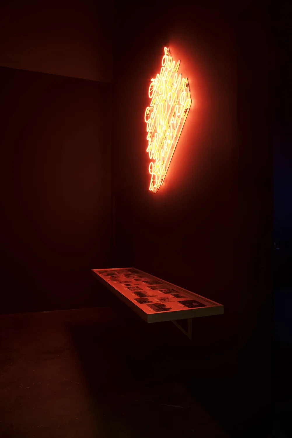 Neon art on the wall spill red light onto vitrine holding documents and photographs below in a dark gallery.