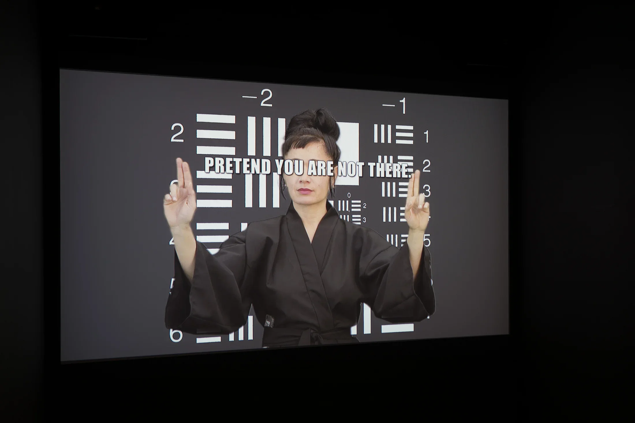 White text “PRETEND YOU ARE NOT THERE” covers the eyes of a woman in black kimono standing before black and white graphic.