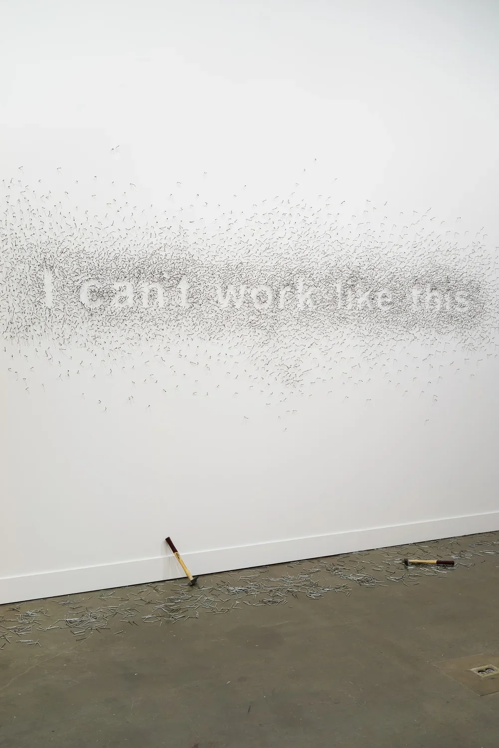 Nails on a white wall form the words “I can't work like this”, on the floor underneath, 2 hammers and scattered nails.