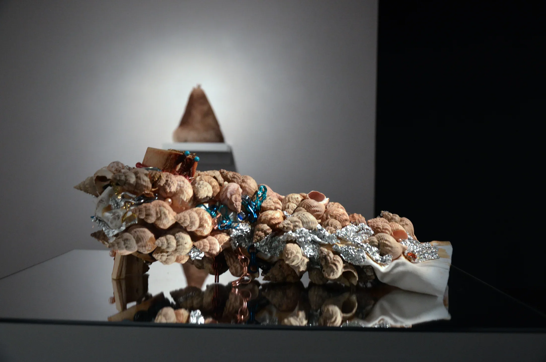 A cascade of tan snail shells on twisted aluminum foil and white fabric rests on a glassy-surface pedestal. 
