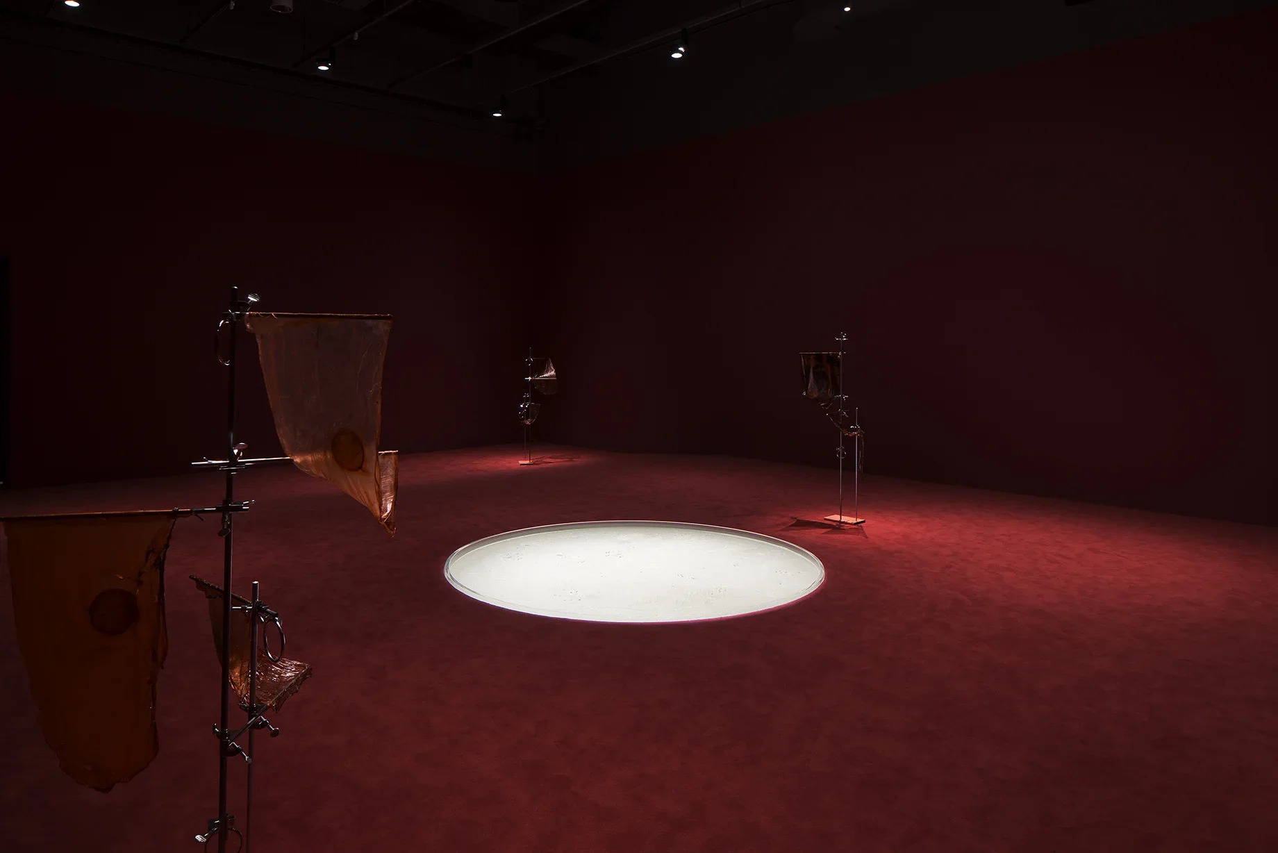 Three stands with draped objects are on the periphery of a room with a white circle in a dark carpeted floor.