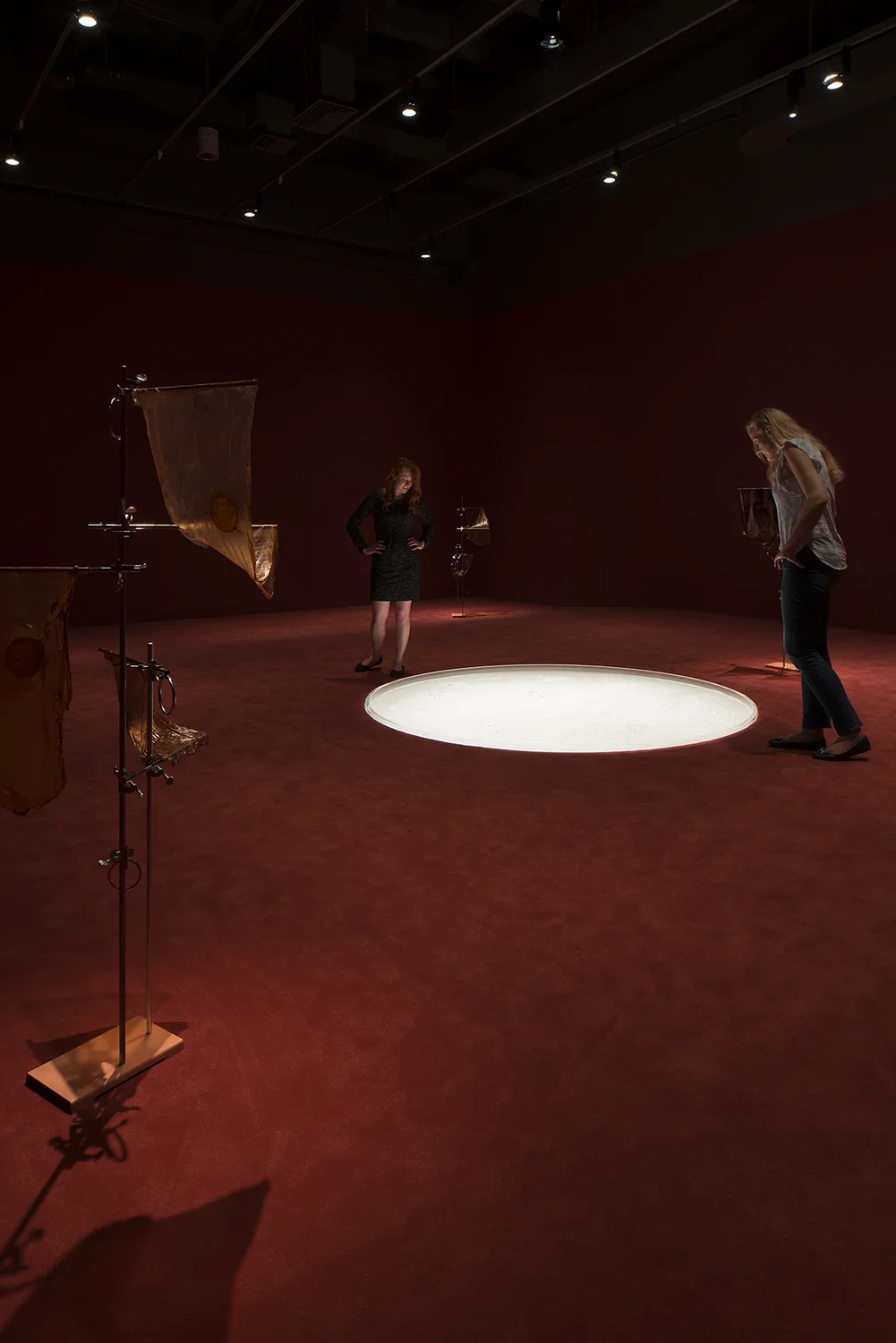 Two guests peer into a white circle embedded in a dark carpeted floor. In the bottom left corner is a stand with a draped object.