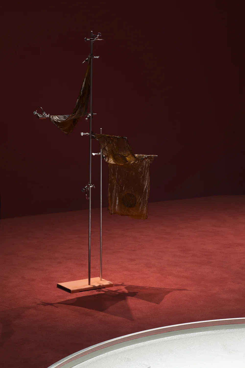 A silver stand with arms akimbo on a wooden base over which are draped diaphanous strands of kombucha.