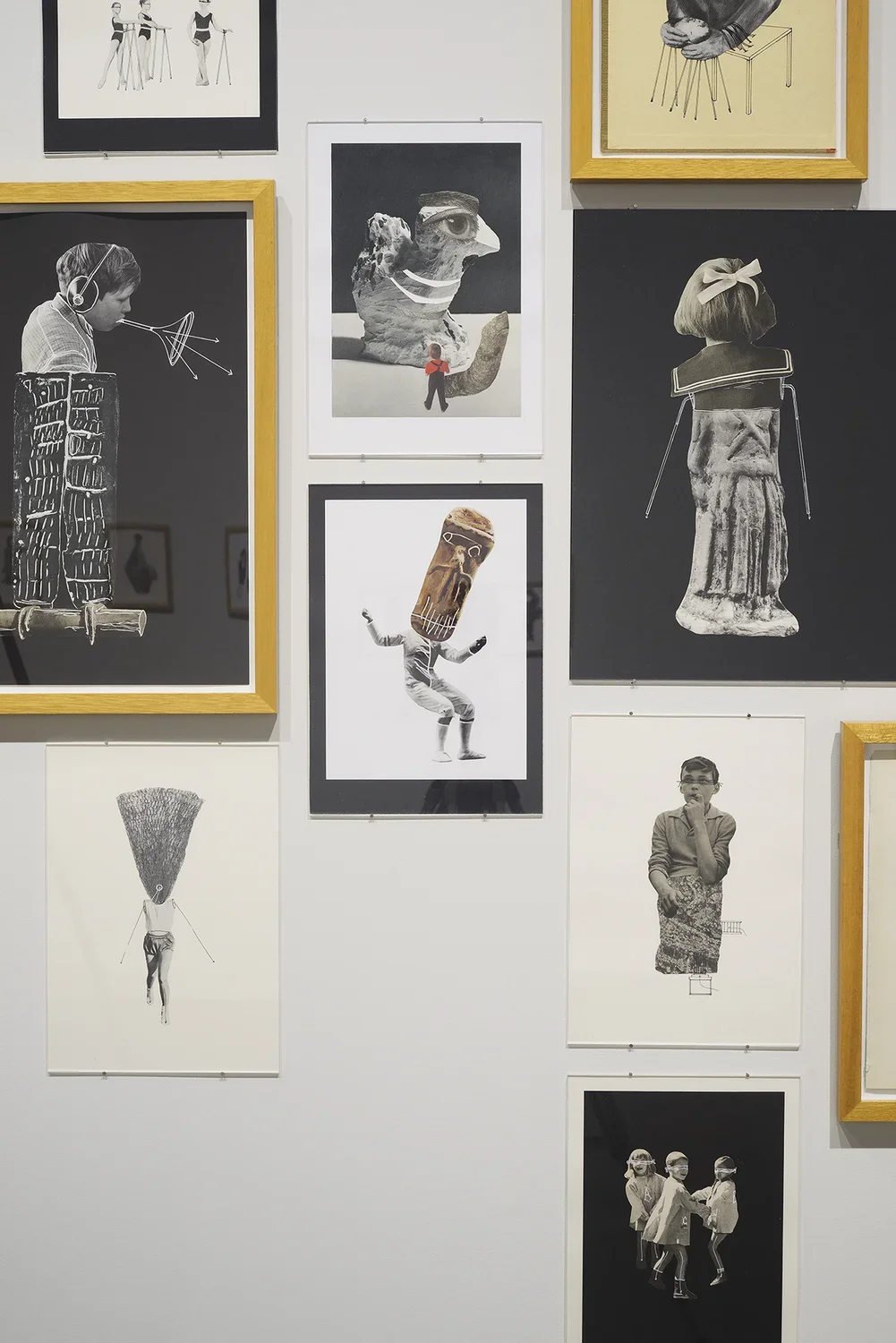 A number of collages of people hang on a wall, some framed and matted, others only matted and rest simply on paper.