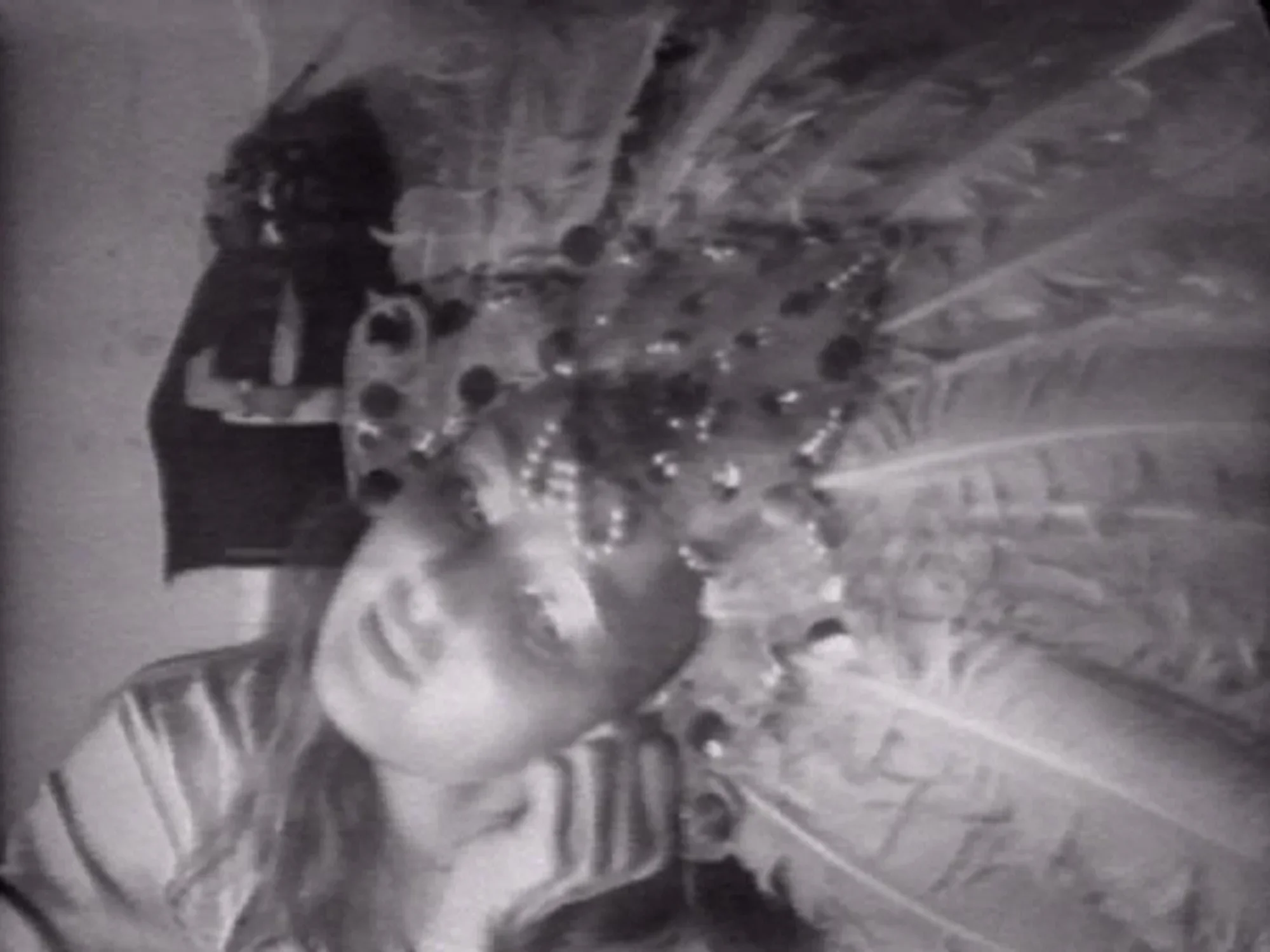 Black and white image of a female stage performer wearing a feather crown headdress with sequins and jewels. Her head is tilted to the side. 