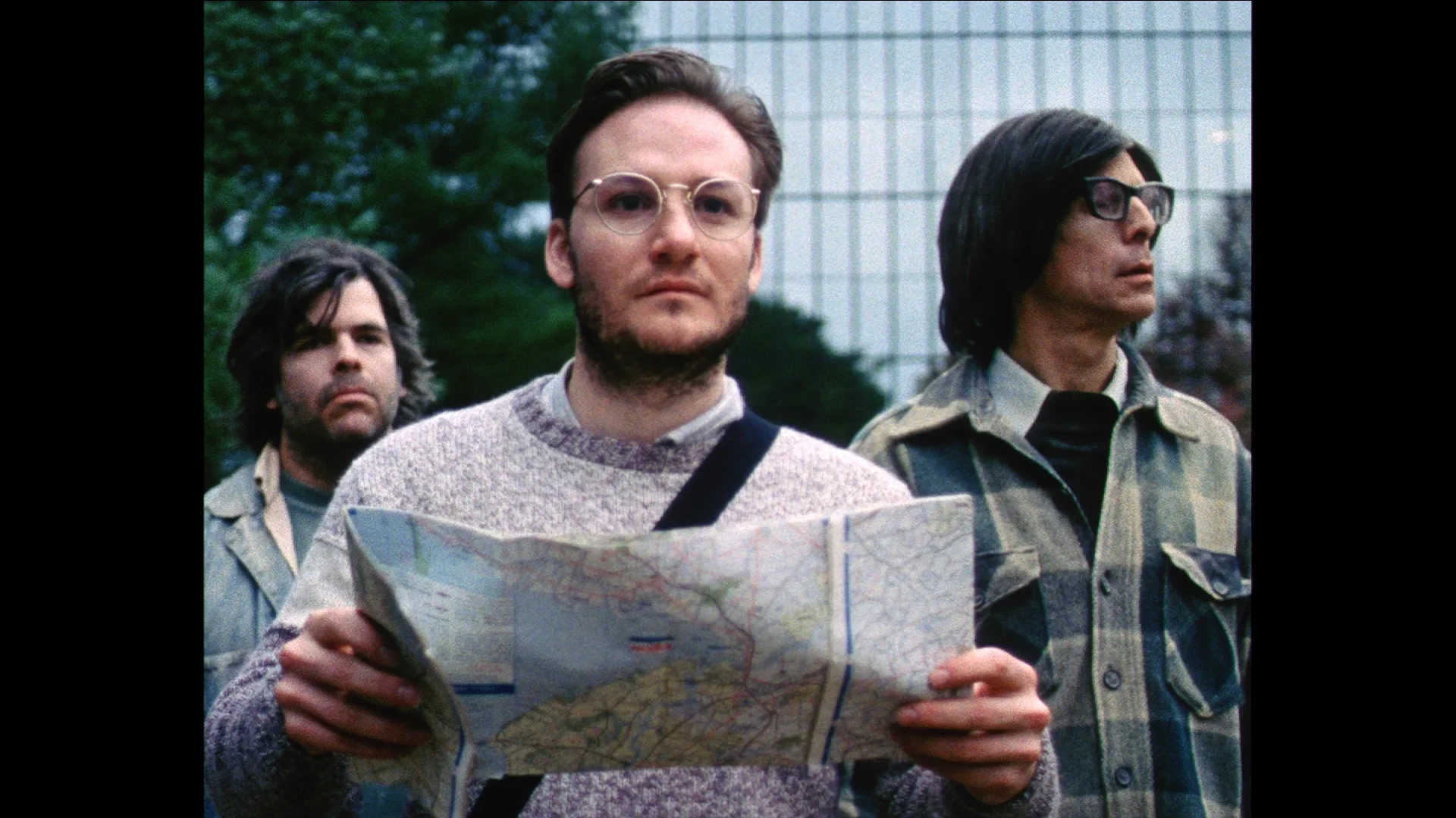 Three men, one in front of the other two, stand outside a mirrored building with landscaping. The man in front holds a map.