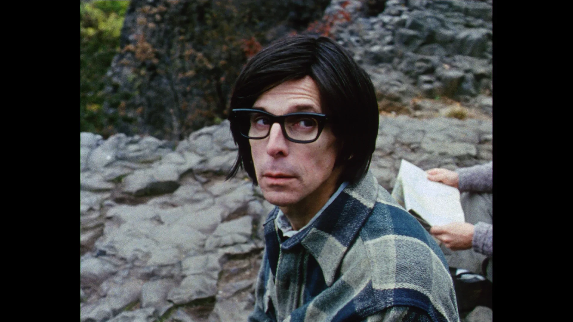 A close up of a person with dark hair and dark glasses wearing a plaid shirt. A set of hands holding a map is to the right.