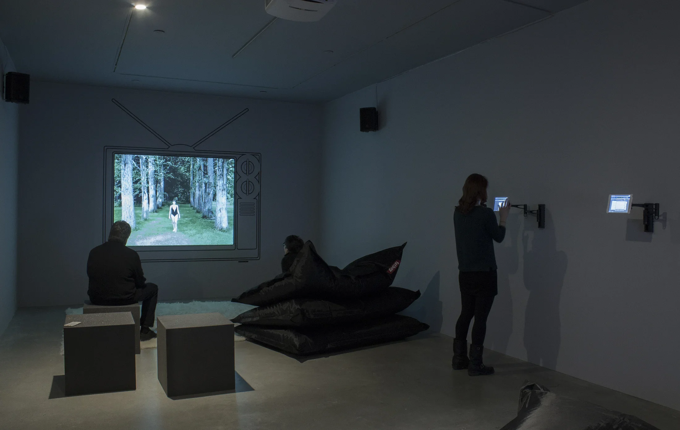 2 visitors watch a projected video framed by the drawing of an old TV, as another visitor interacts with a wall-mounted iPad.