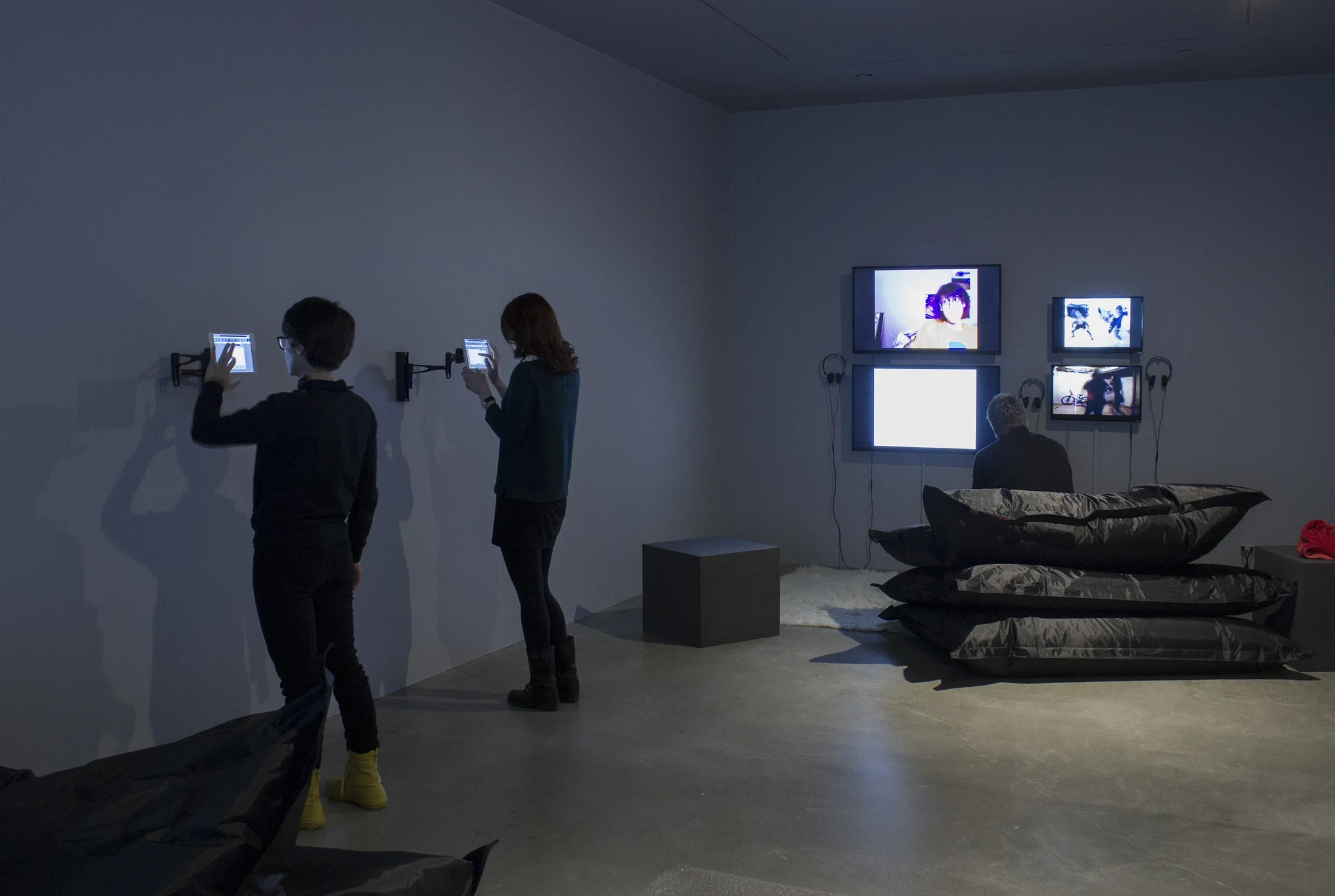 2 visitors interact with wall-mounted iPads while a visitor sits on beanbags watching 4 wall-mounted monitors.