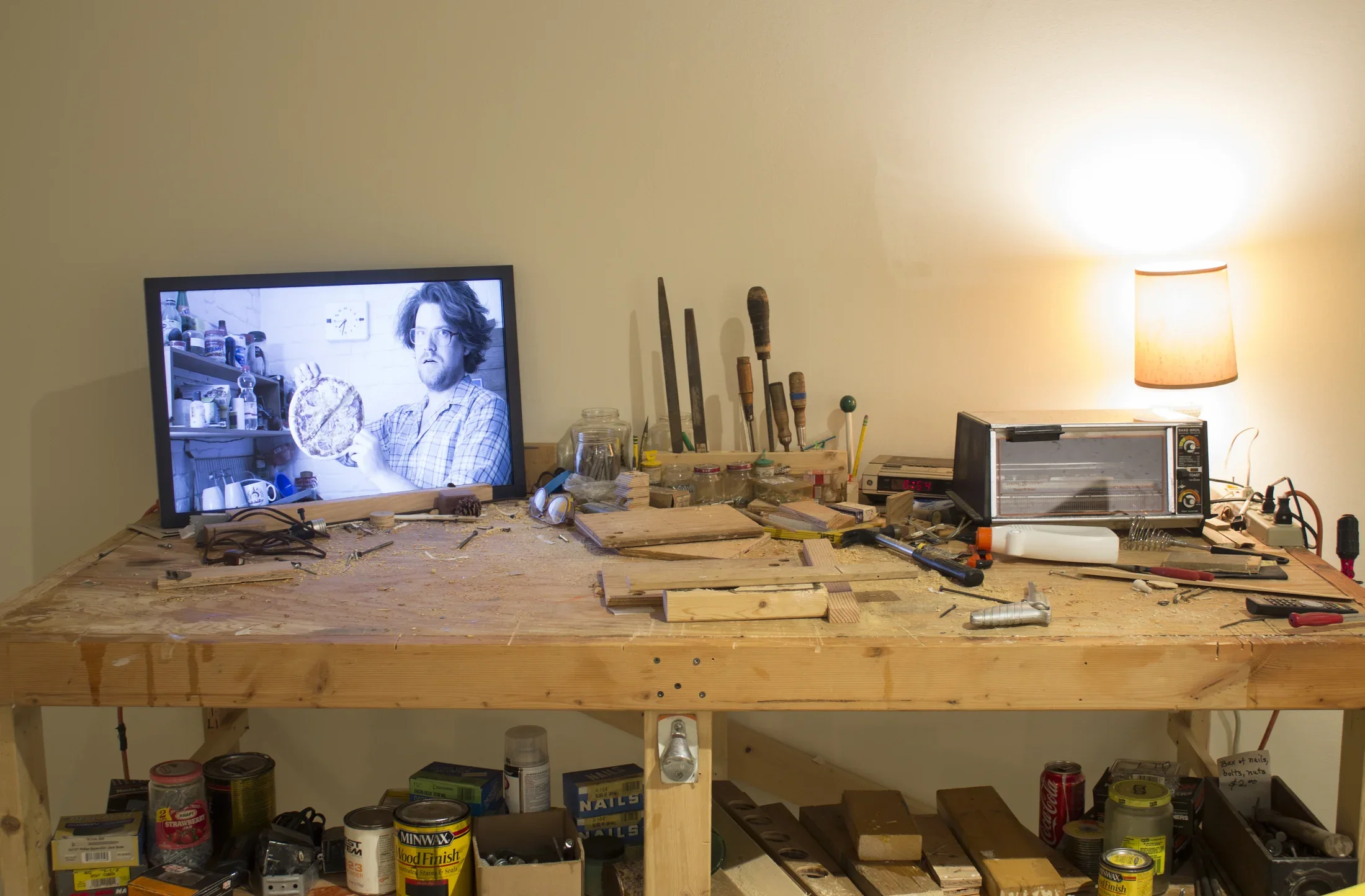 Wood work bench covered in scraps, tools, a toaster oven, a lamp, and a monitor with an image of the artist holding a pizza