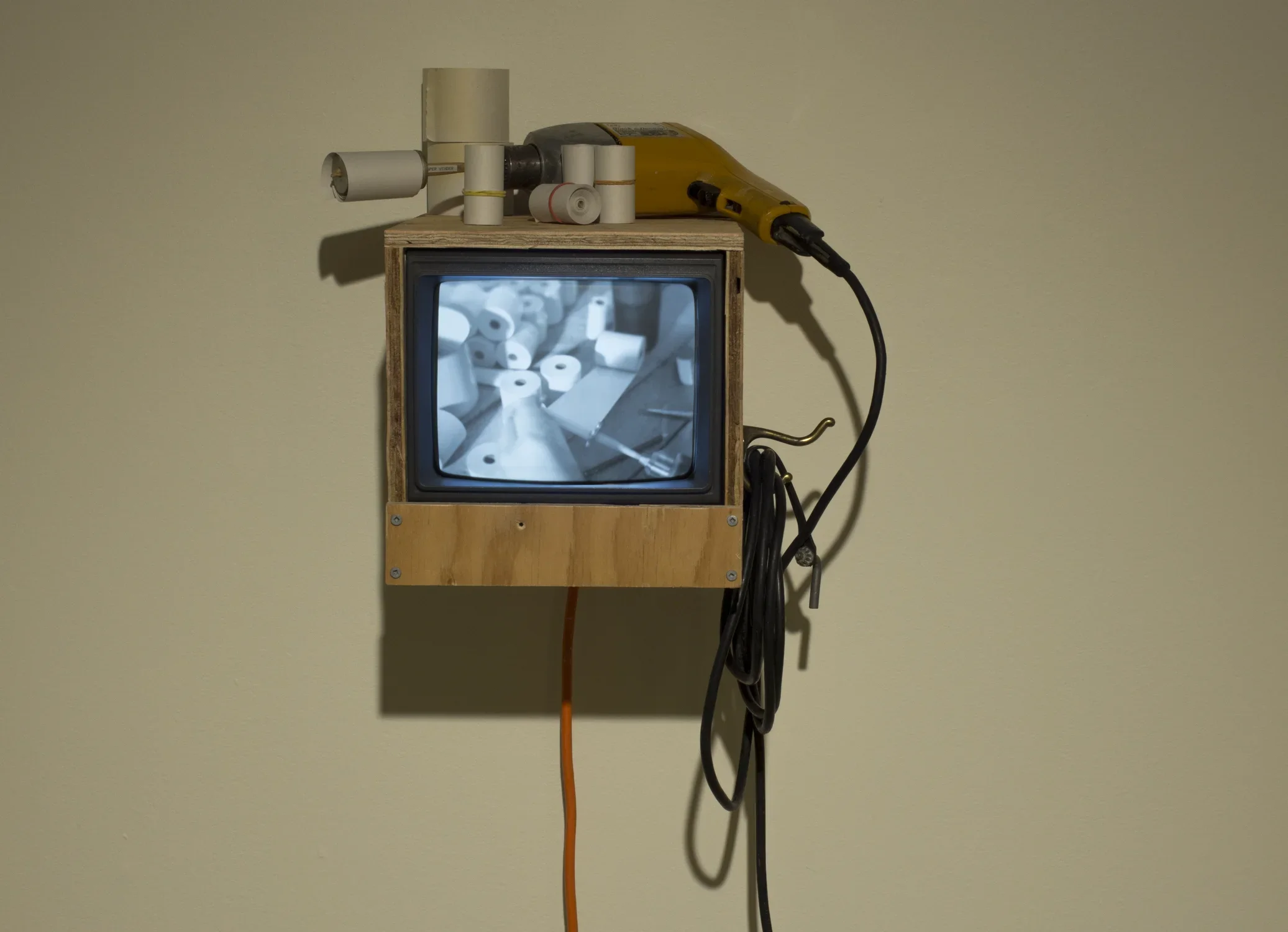A monitor with an image of paper rolls hangs on a wall in a wood frame, an electric drill and rolls of paper sit on top
