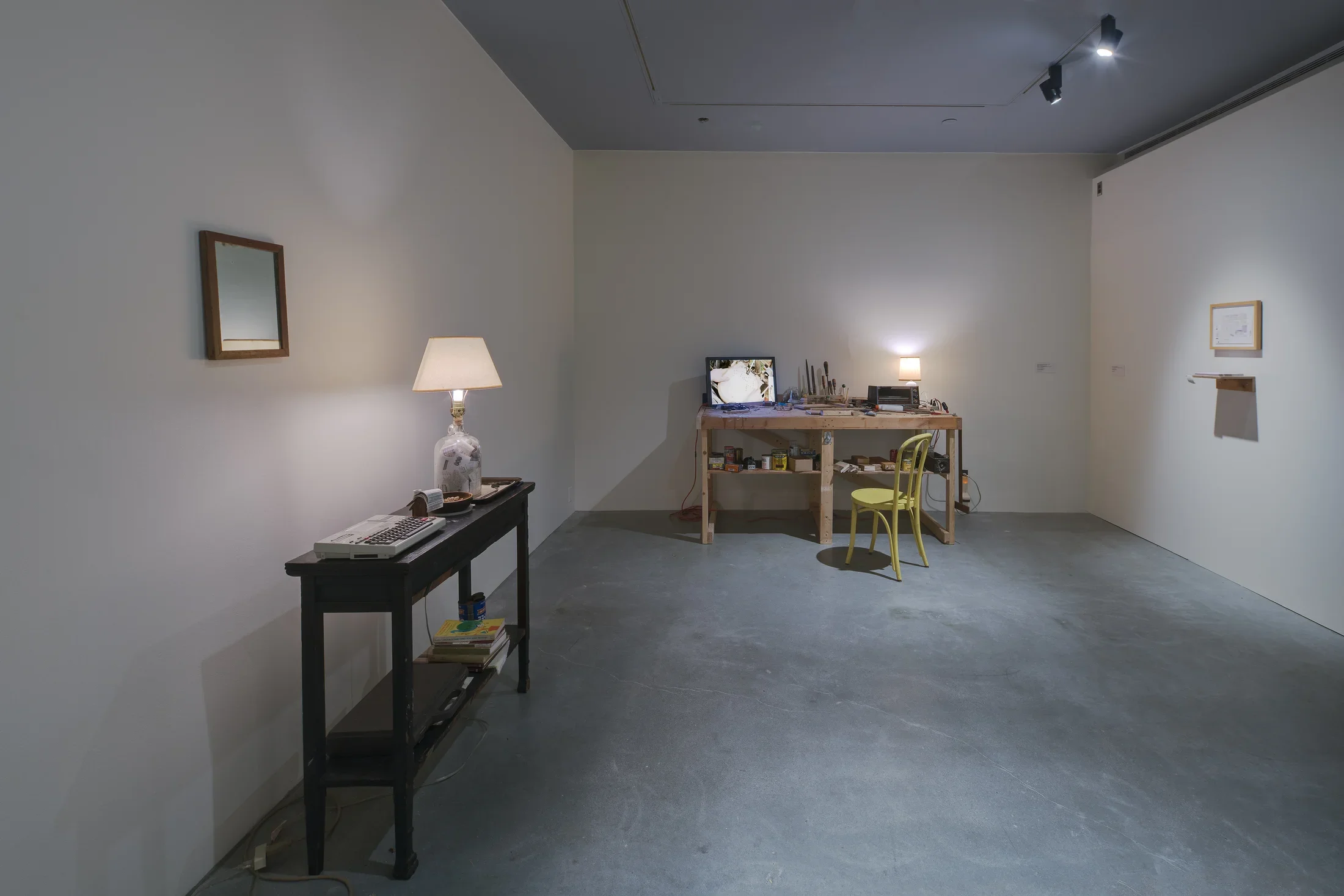 A space with workbench, lamp and chair in back, a picture and shelf on the right, a mirror and table with a lamp on the left