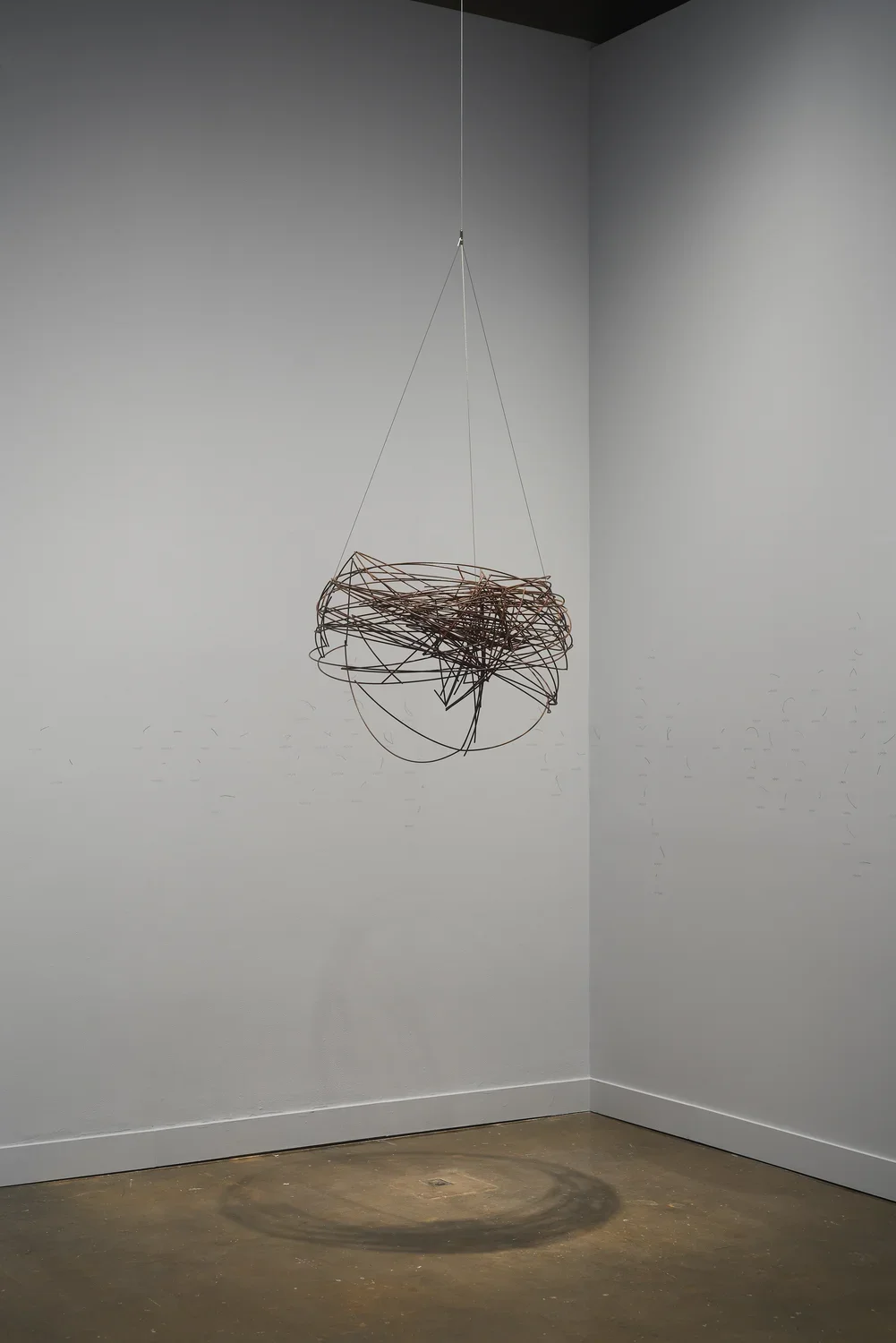 A spherical, brown-bronze-color metal-wire sculpture hangs from the ceiling before white walls in a corner of the gallery.