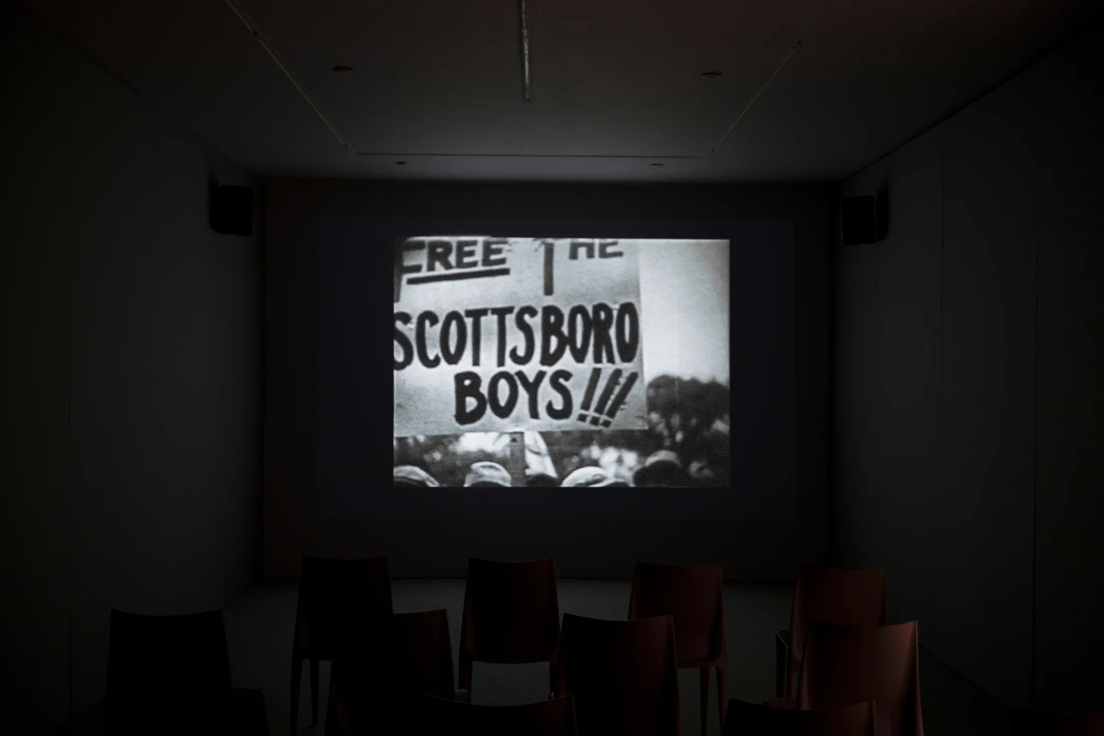 In the center of a dark room, a black and white video displays a protest sign that says “Free the Scottsboro Boys”.