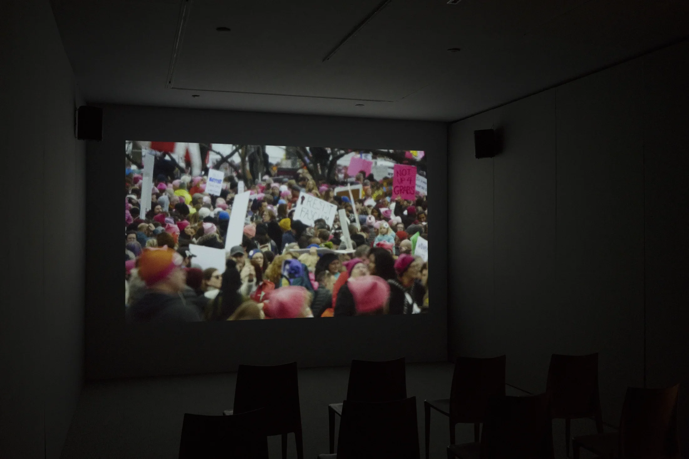 A colorful video scene of a large crowd of protesters, many holding signs, plays on a screen in a dark room.