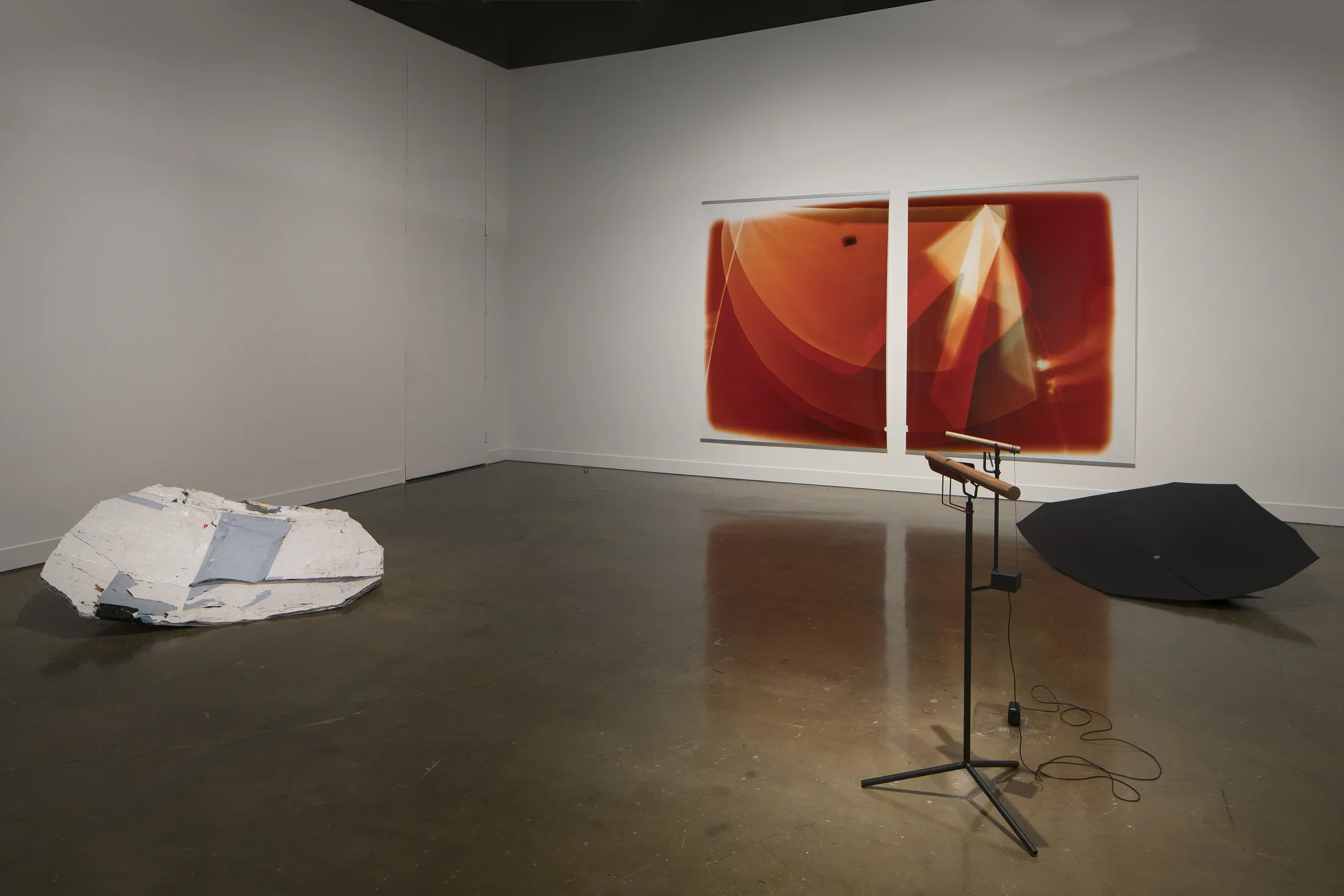 Two orange and white prints on a wall, a black and a white shield sculpture on the floor, a wood sound piece on a stand