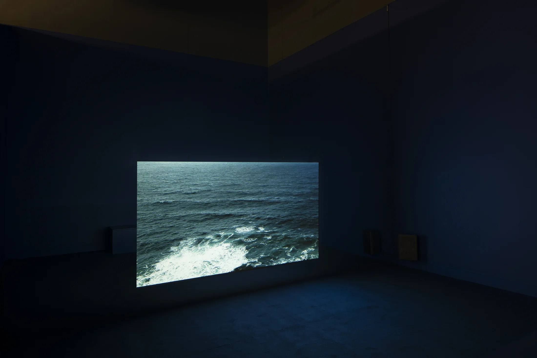 A pitch black space with a projection of a dark blue body of water with ripples and a white, crashing wave