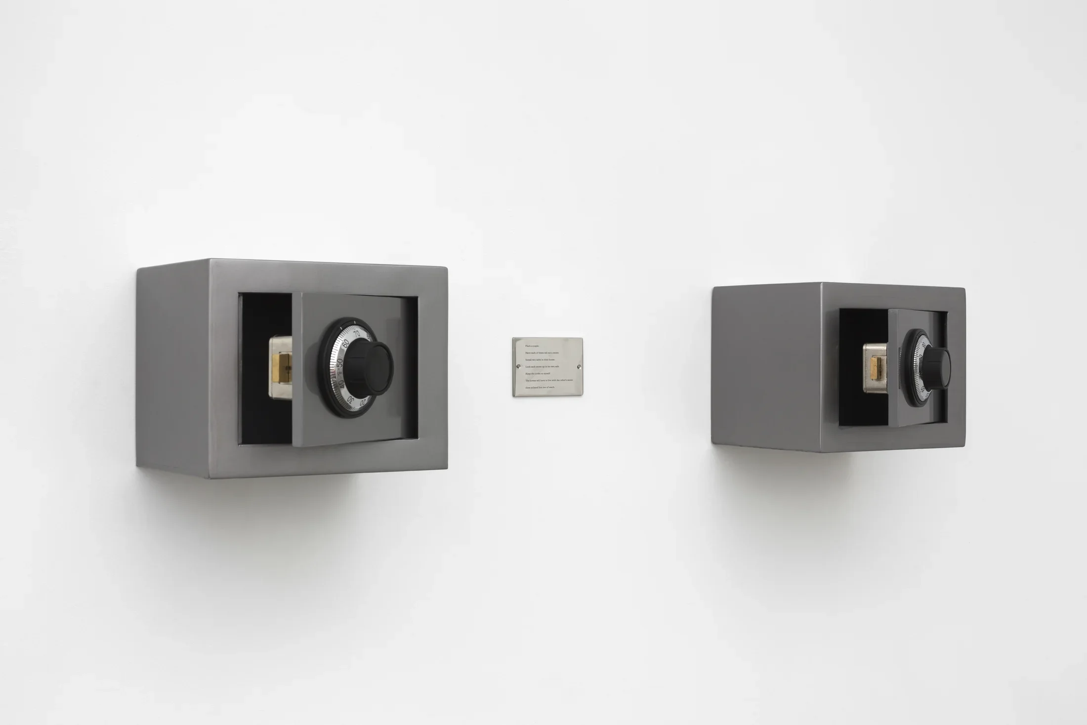 Two small rectangular safes, both partially open and secured on a white wall, an engraved plaque centered between them
