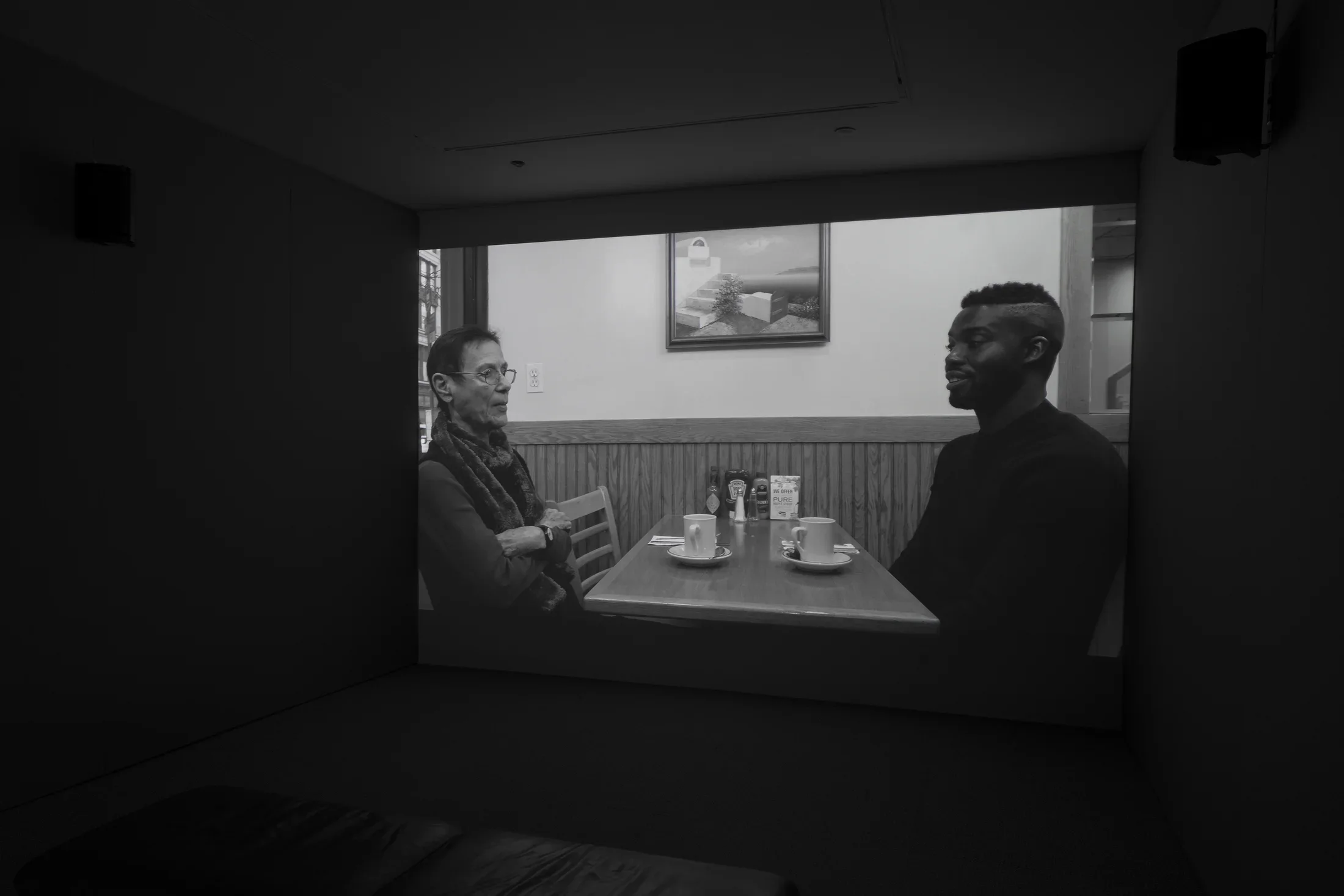 Black and white video projection of a woman and a man in conversation, sitting opposite at a cafe table set with coffee.