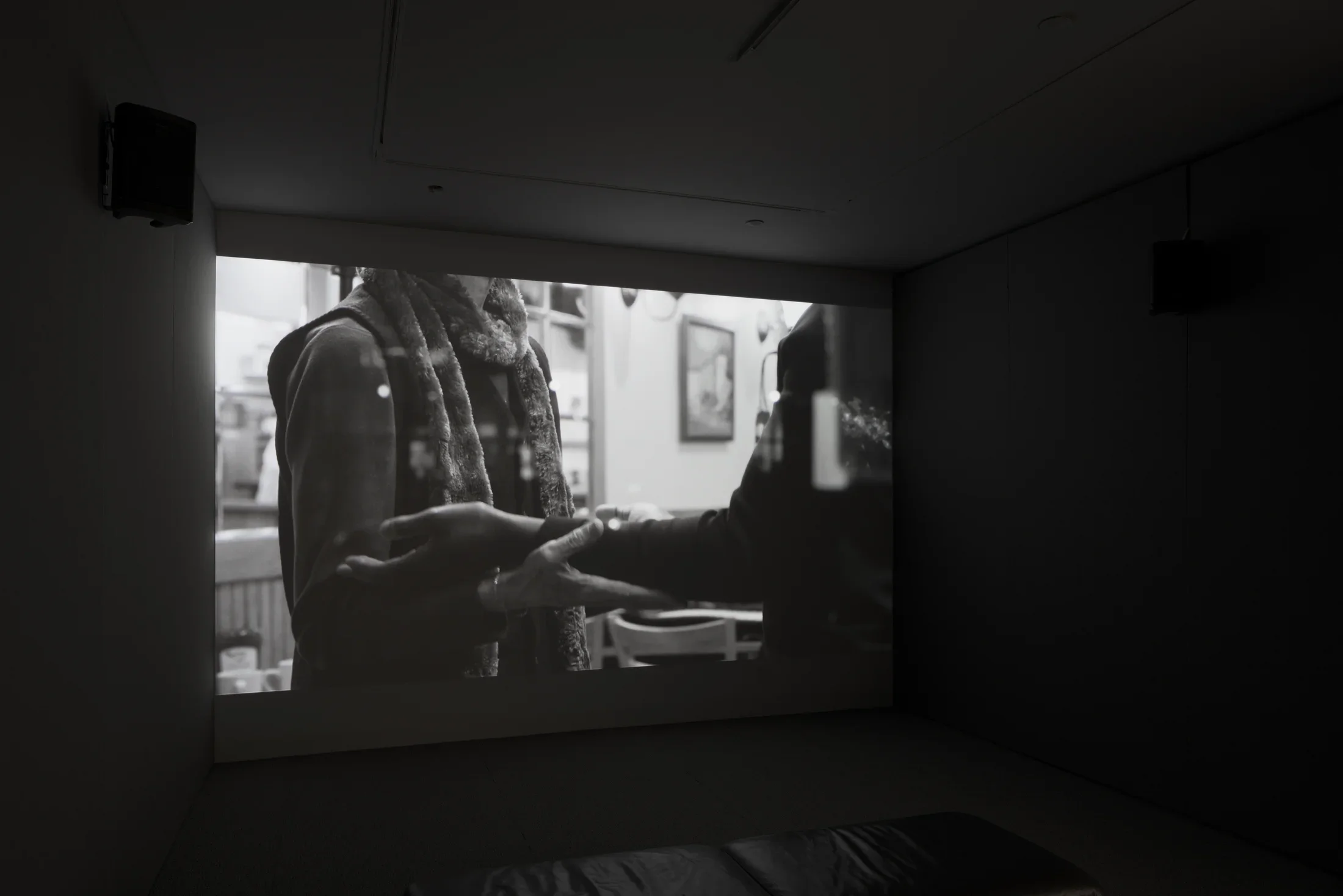 Dark room with video projection of a person's torso, arms bent at right angle, palms up, supporting the arms of another.