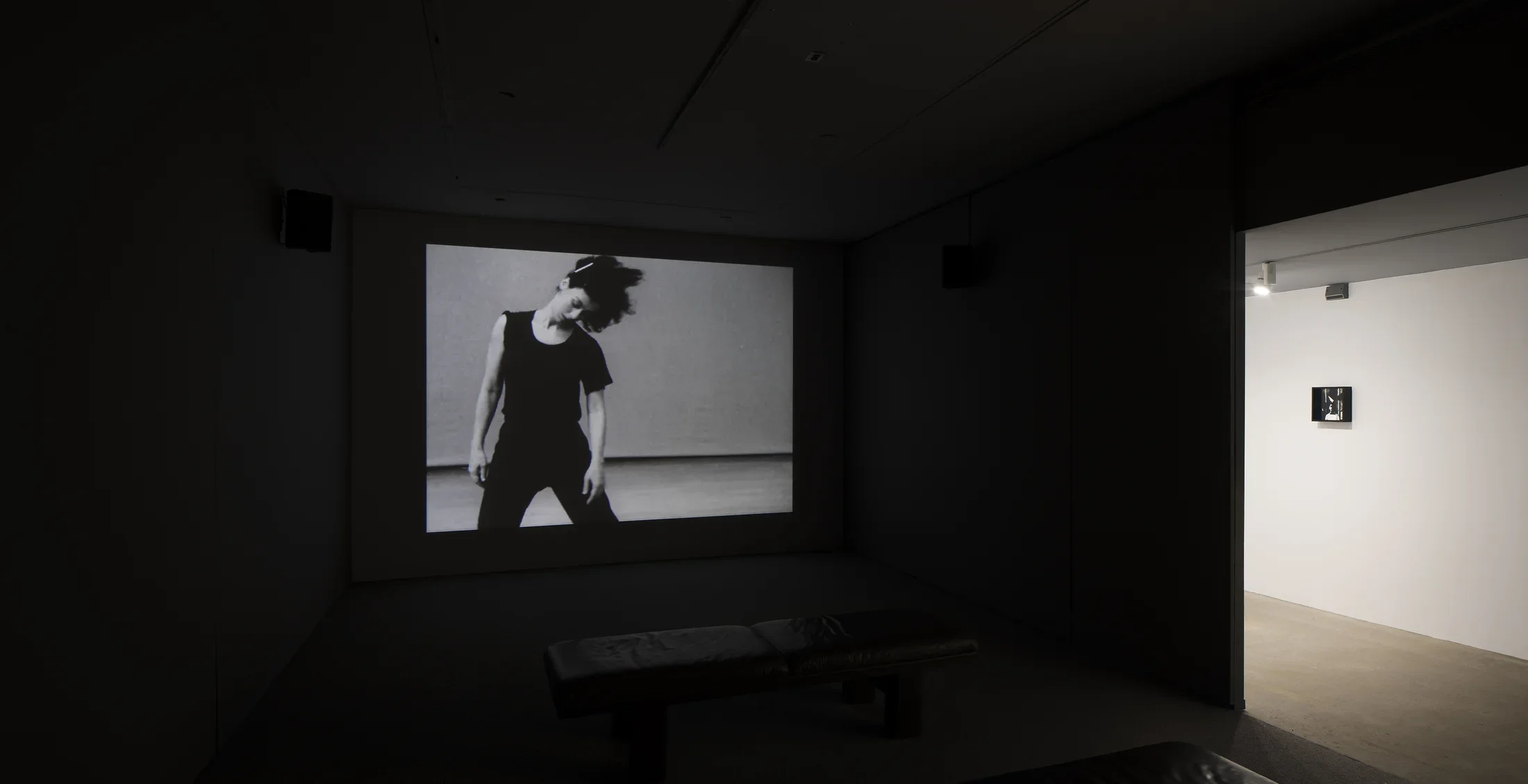 A dancer in motion is projected in a dark room. An opening leads to a bright hall where one small work hangs.