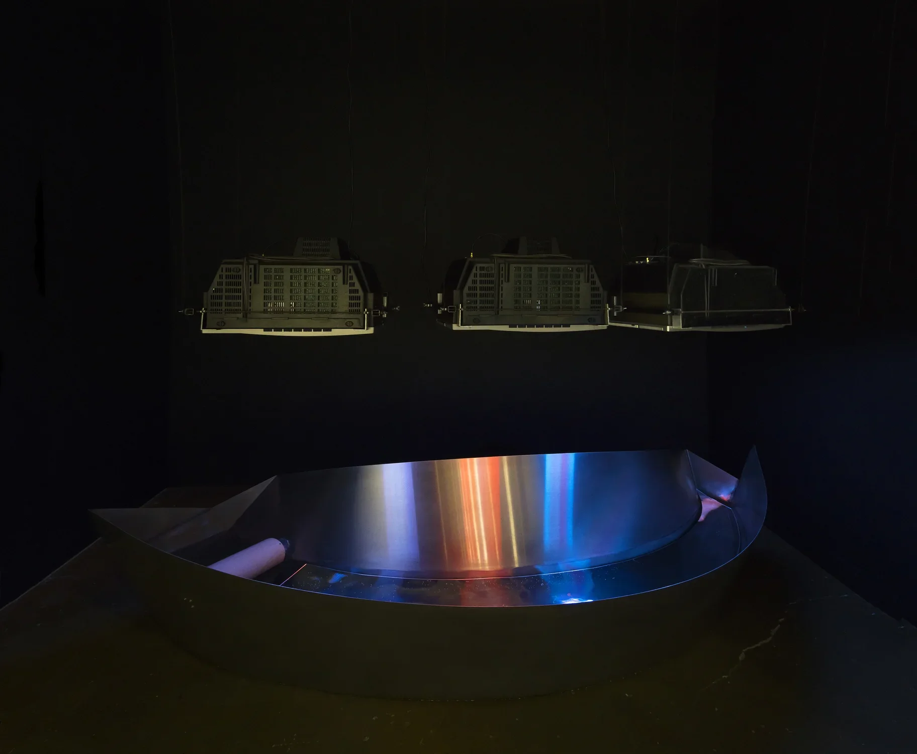A curved, steel trough reflects colored images from 3 CRT monitors, suspended face-down above it, in a dark space.