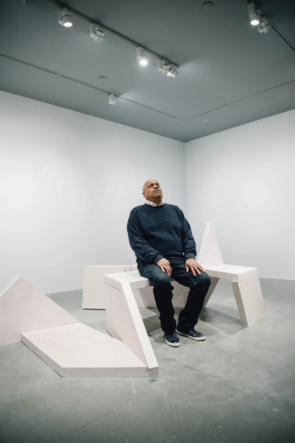 A man seated on a bench sculpture leans back slightly, his head tilted up with eyes closed, hands on his knees.
