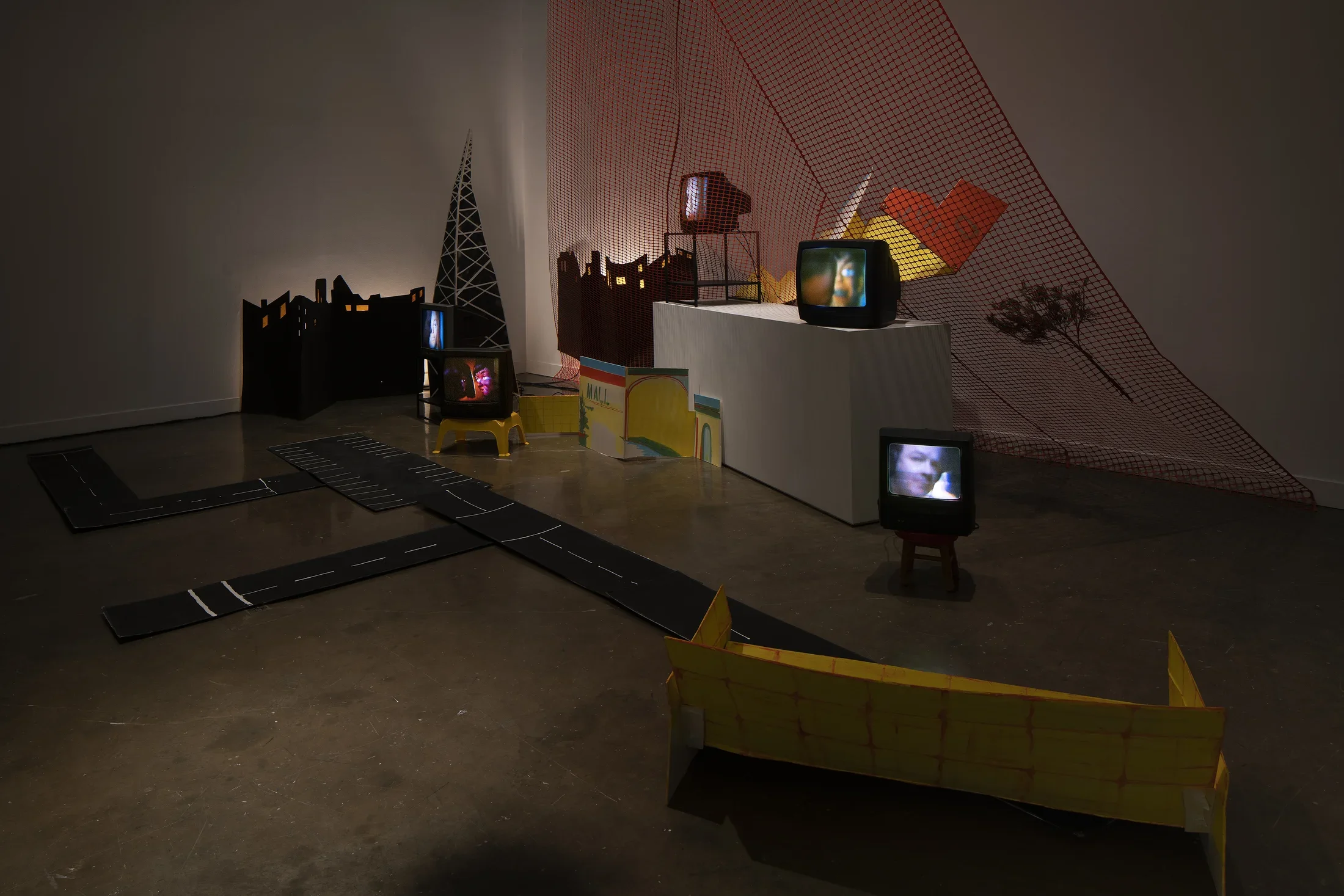 Dark image of five tv monitors on the floor and on stands, with foam core and cardboard elements, as well as plastic fencing.
