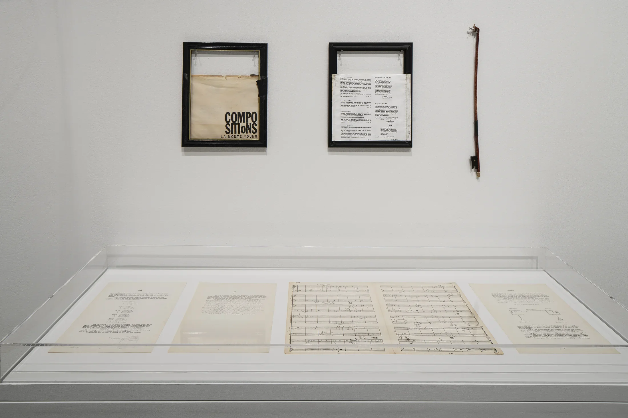 Two framed objects and a half-sized bow hang on a wall behind a glass vitrine containing a musical score and other printed pages.