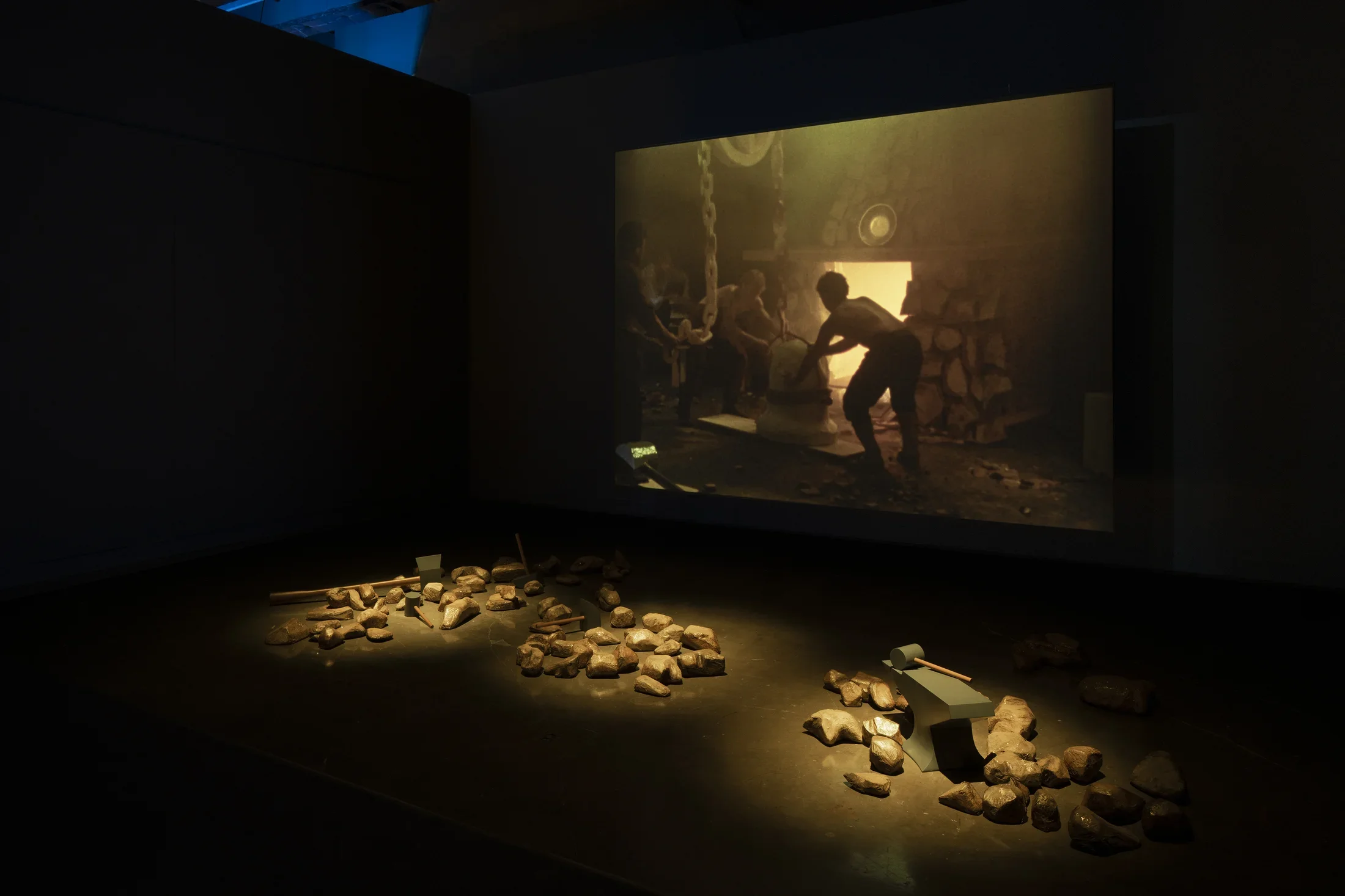 Three piles of gold-painted rocks and mallets are grouped in front of a screen on which an image of a man working in a forge.