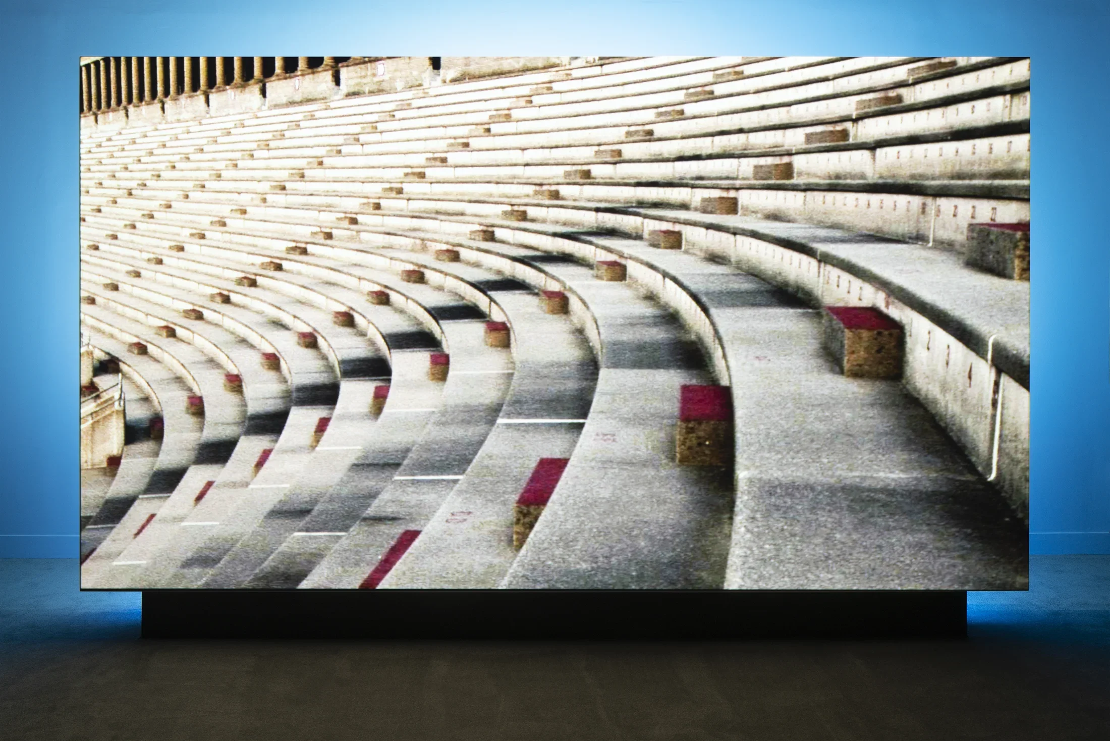 A large screen with the projected image of convex concrete stadium seating stands in the Gallery, backlit by a blue light.