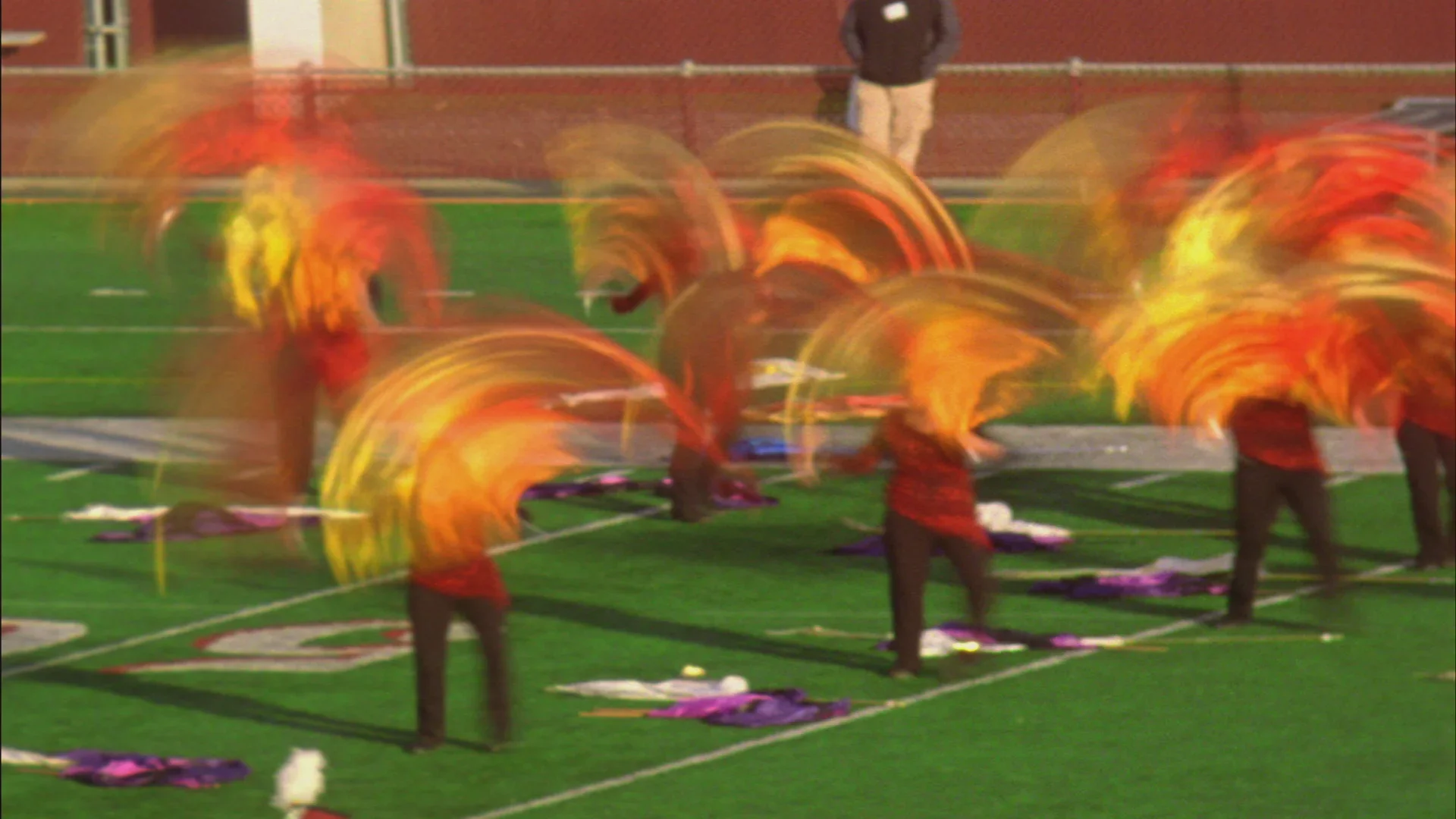 Video still of a drum corps wearing red and black, waving colorful flags on the 50 yard line of a football stadium.