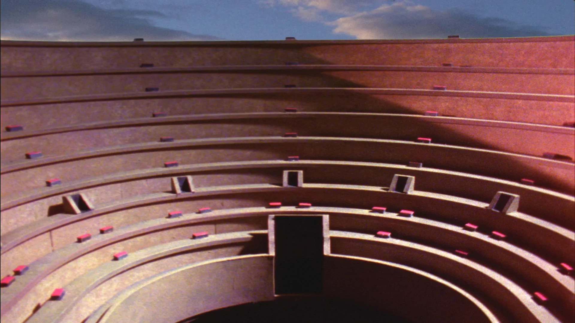 Video still of upper reaches of the convex seating of an open-air stadium. Blue sky and clouds visible at top of image.