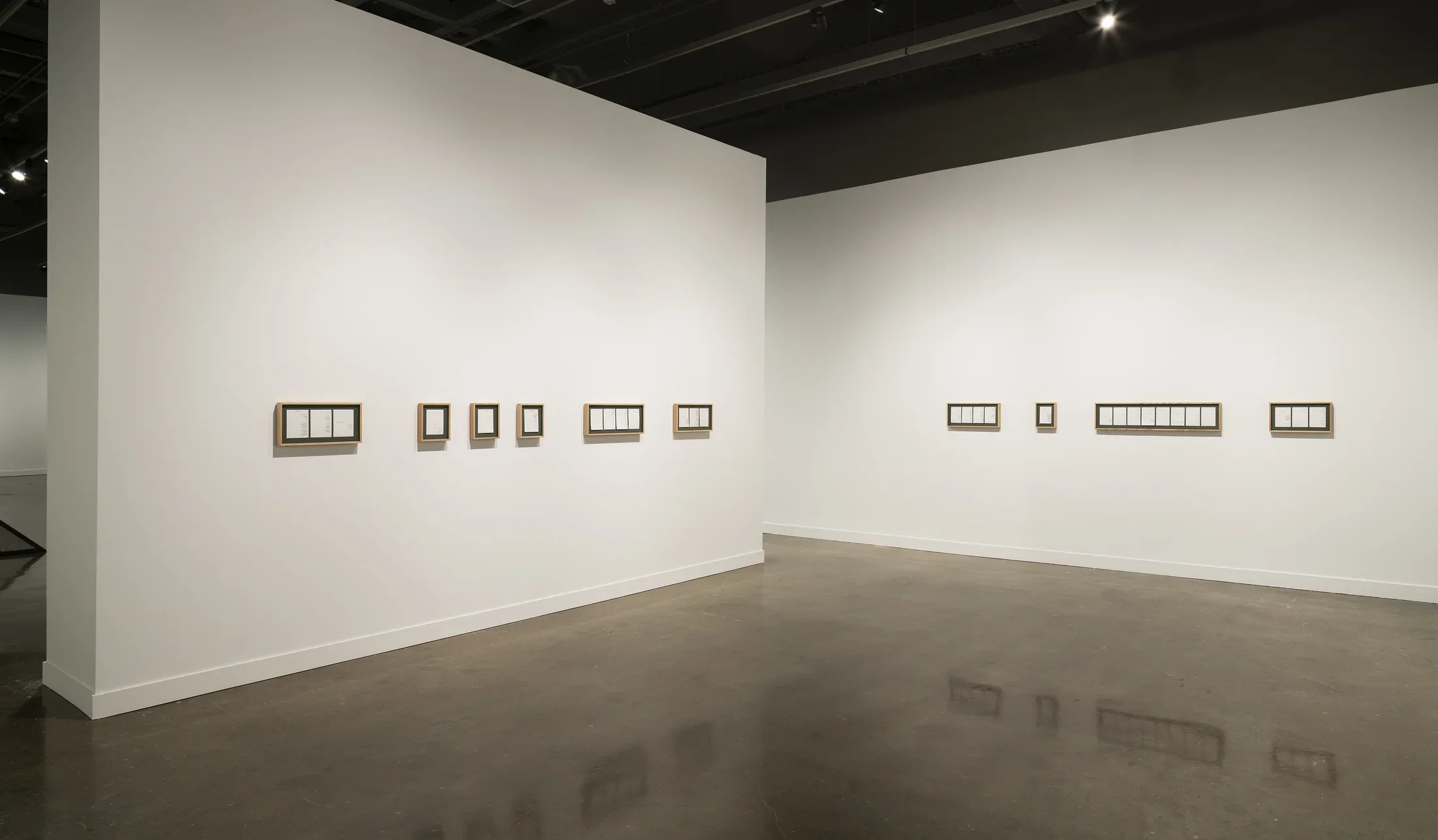 Nine horizontal frames with dark matting and white pieces of paper hang from two nearly bisecting white walls.