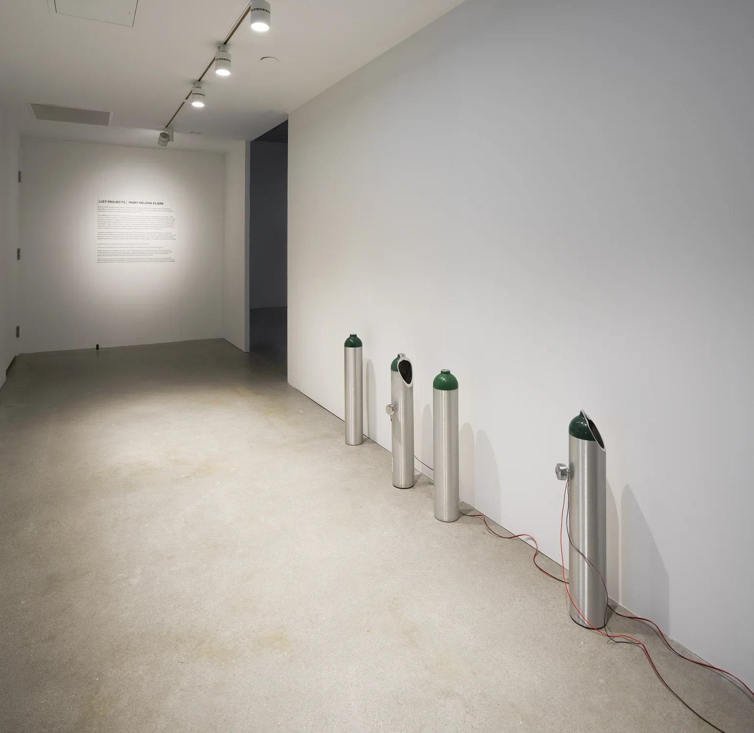 On the right side of a hallway sit 4 cylindrical tanks with painted green tops, some with holes and wires. Text on back wall.