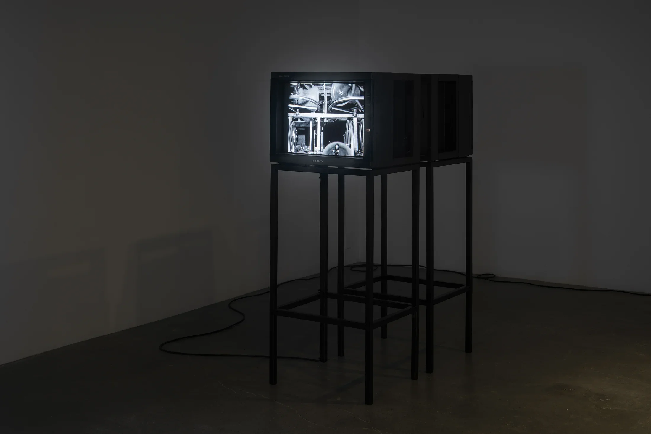 Two video monitors on stands face in opposite directions in a darkened room. A black and white image is visible on one monitor.